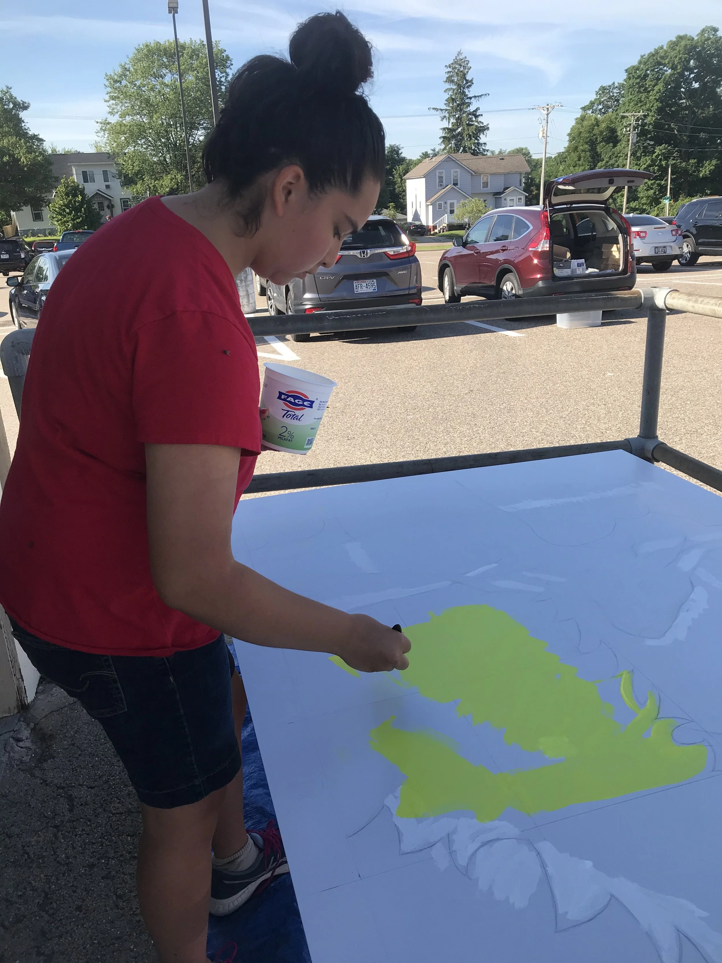 A young woman wearing a red t-shirt and black shorts is painting a large map of a green landmass on a white canvas outdoors in a parking lot with cars and houses in the background.