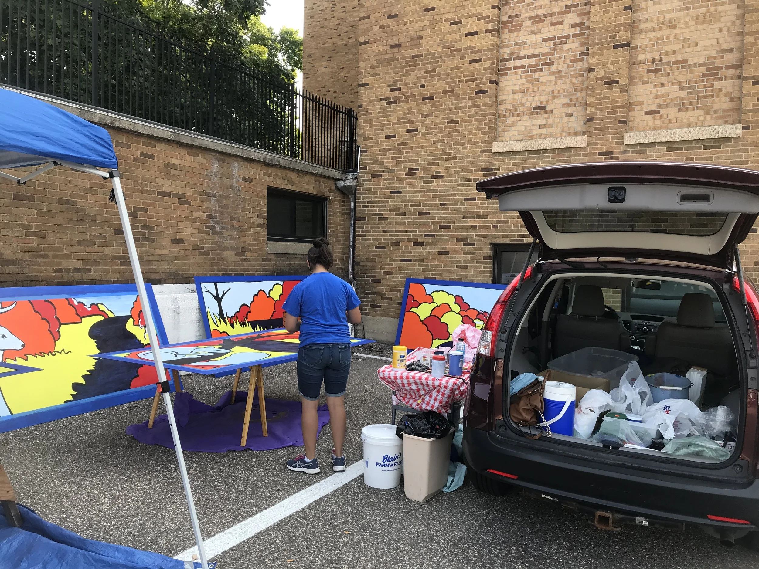Woman painting colorful outdoor murals on large panels set up next to her car in a parking lot.