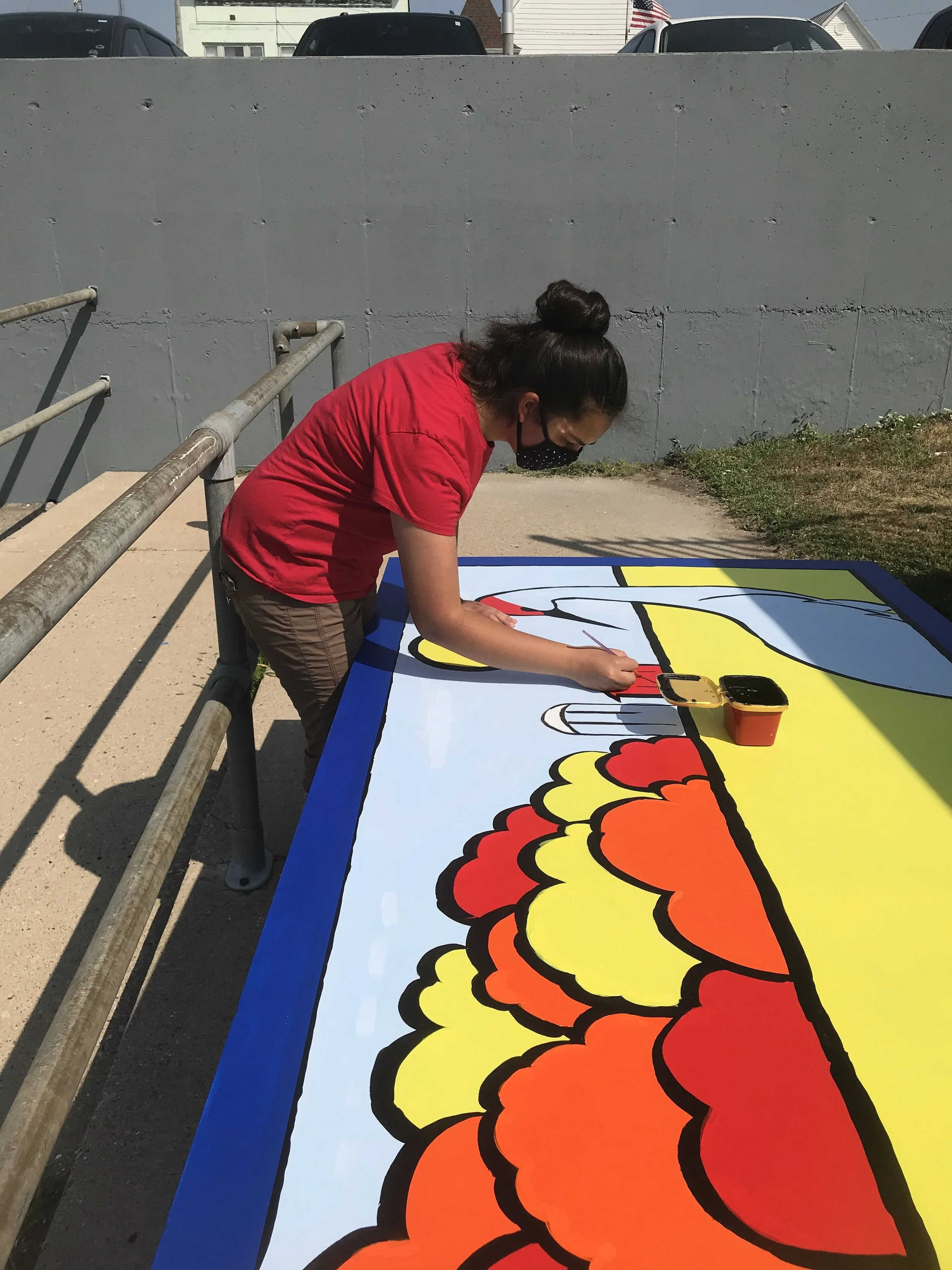 A person painting a colorful mural outdoors on a large flat surface, wearing a red shirt and a face mask.