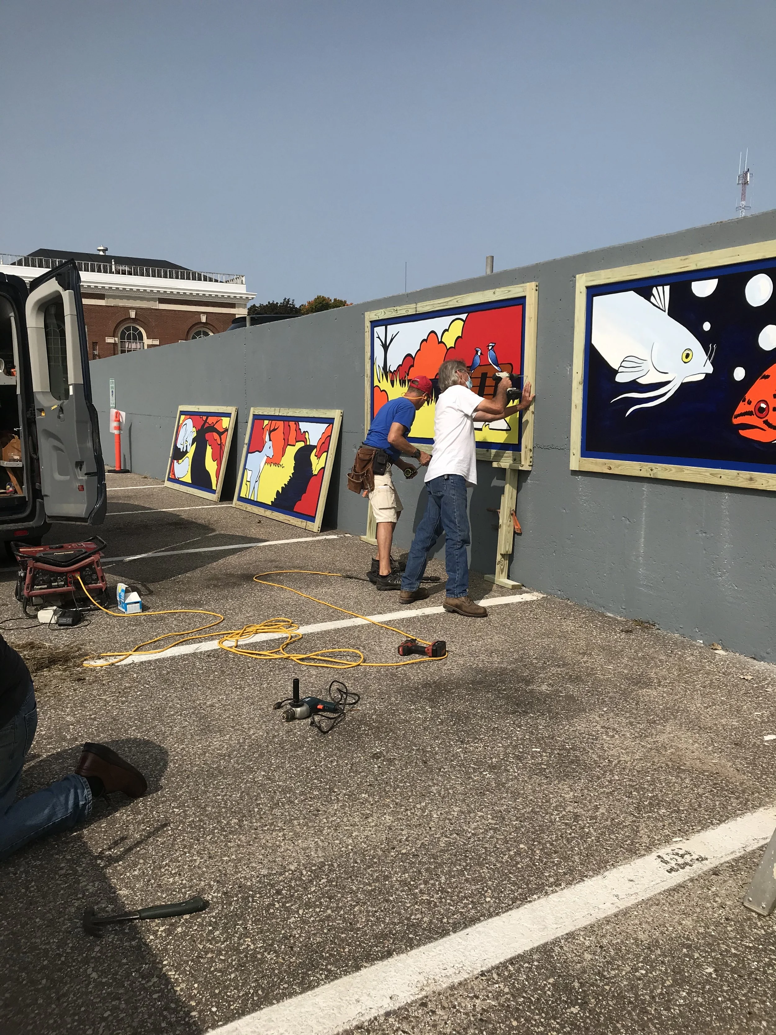 Two artists working on colorful paintings of animals, including fish, in a parking lot, with a gray wall and various tools on the ground.