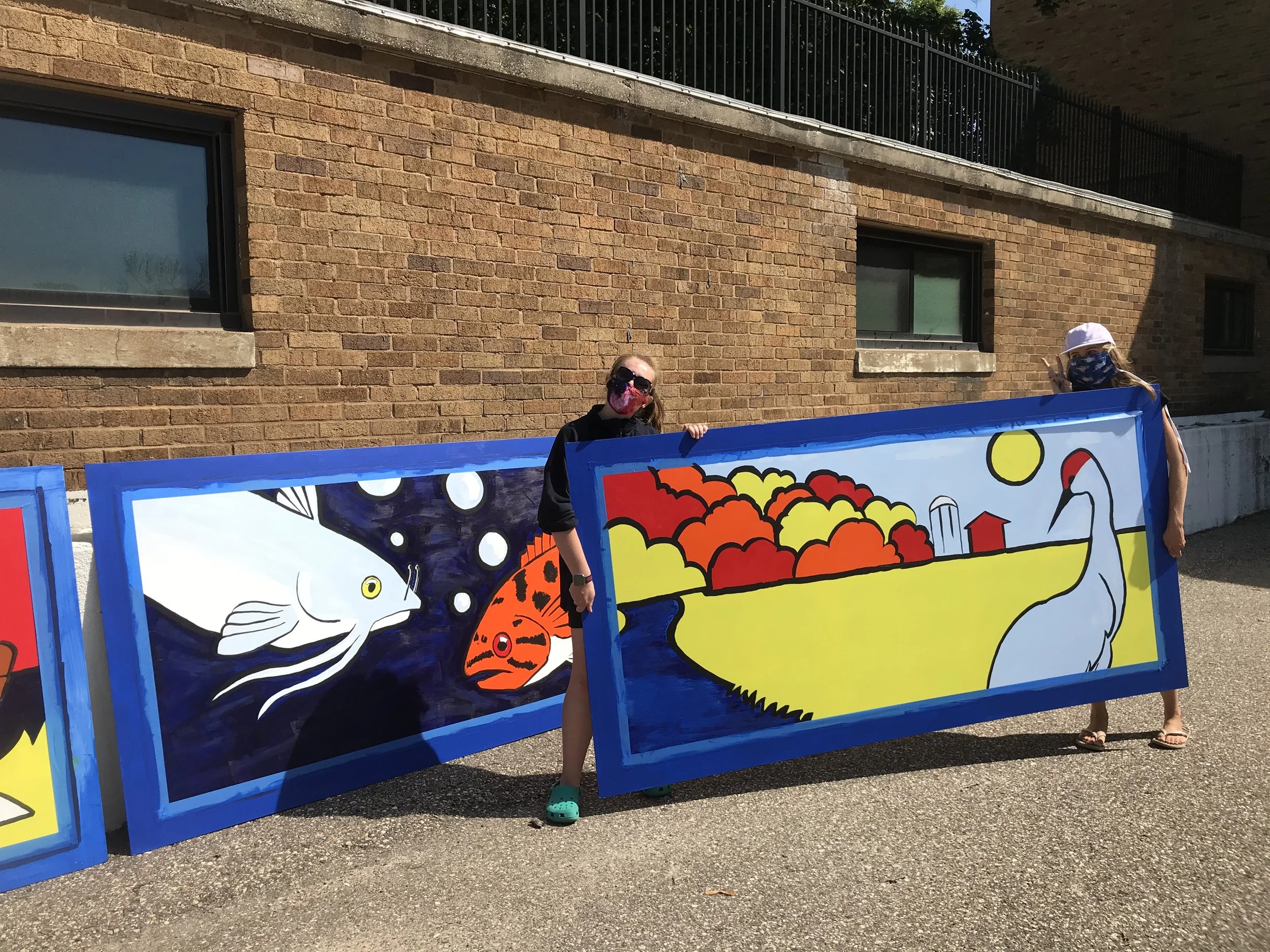 Two people wearing masks holding colorful paintings of various animals and scenery outdoors near a brick building.