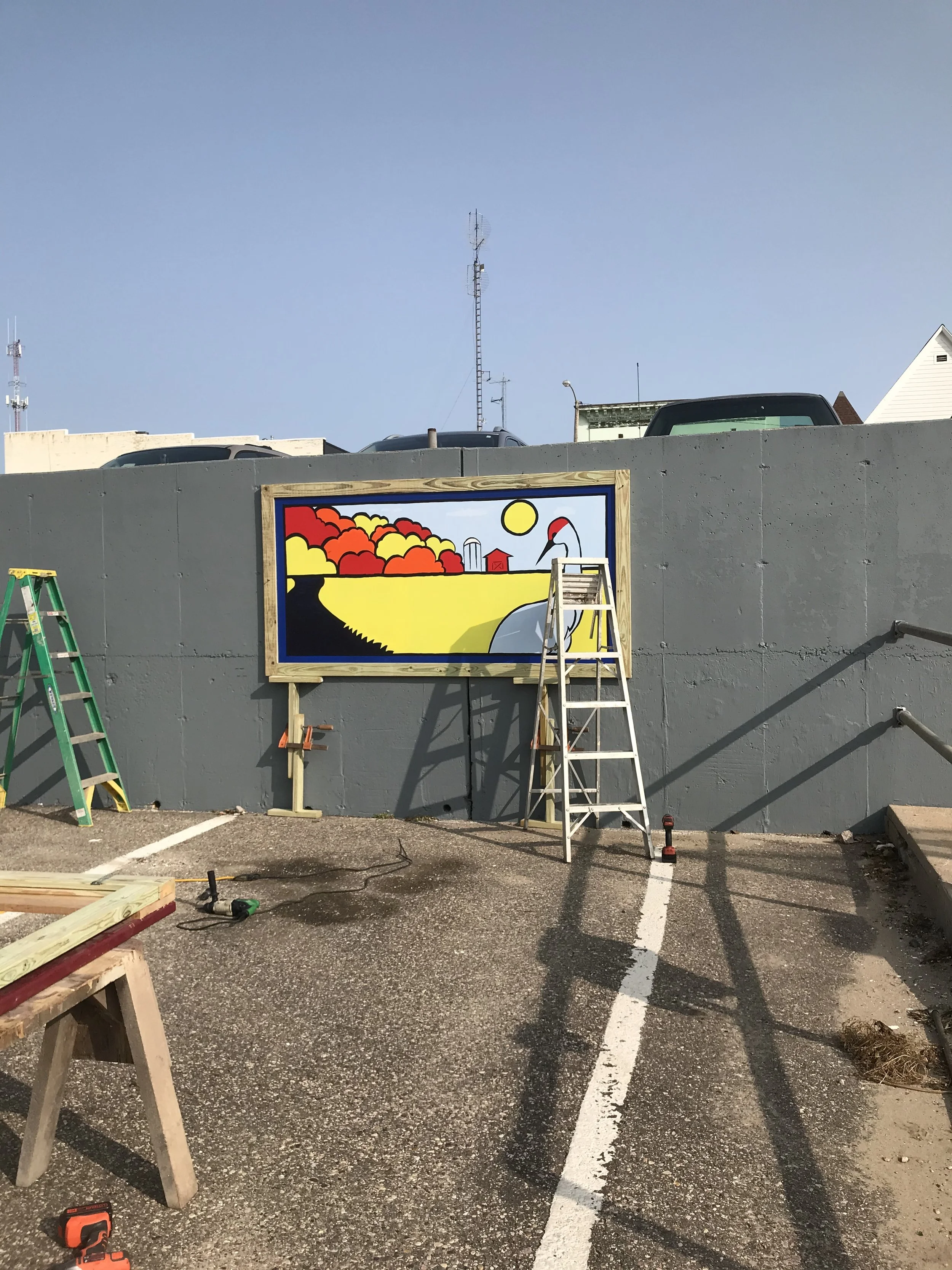 A colorful mural of a landscape with a large bird, likely a heron, on a wall under construction, with ladders and construction tools nearby, set in a parking lot.