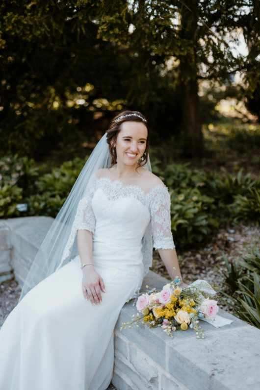 Bride in a white lace wedding dress sitting on a stone ledge outdoors with a bouquet of pink and yellow flowers beside her.