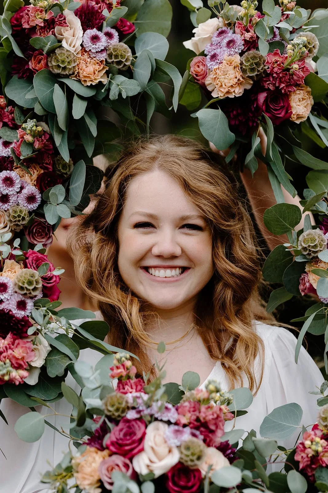 A woman with curly red hair smiling through a floral wreath made of colorful flowers and green leaves.
