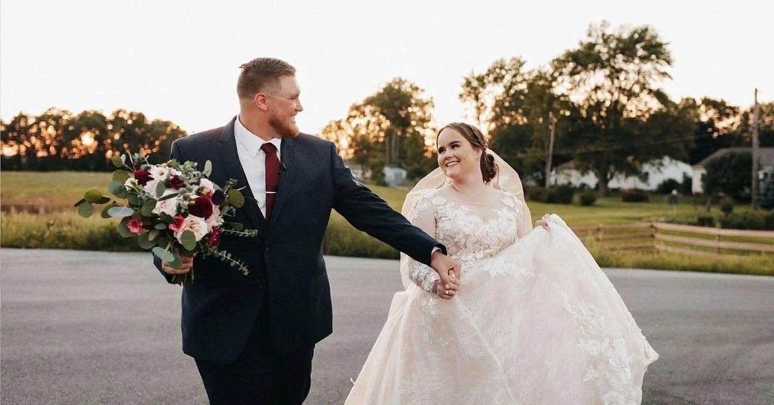 A couple on their wedding day walking hand in hand outdoors during sunset. The groom is dressed in a black suit with a red tie, holding a bouquet of flowers, while the bride wears a white lace wedding gown, lifting her dress slightly and smiling at t