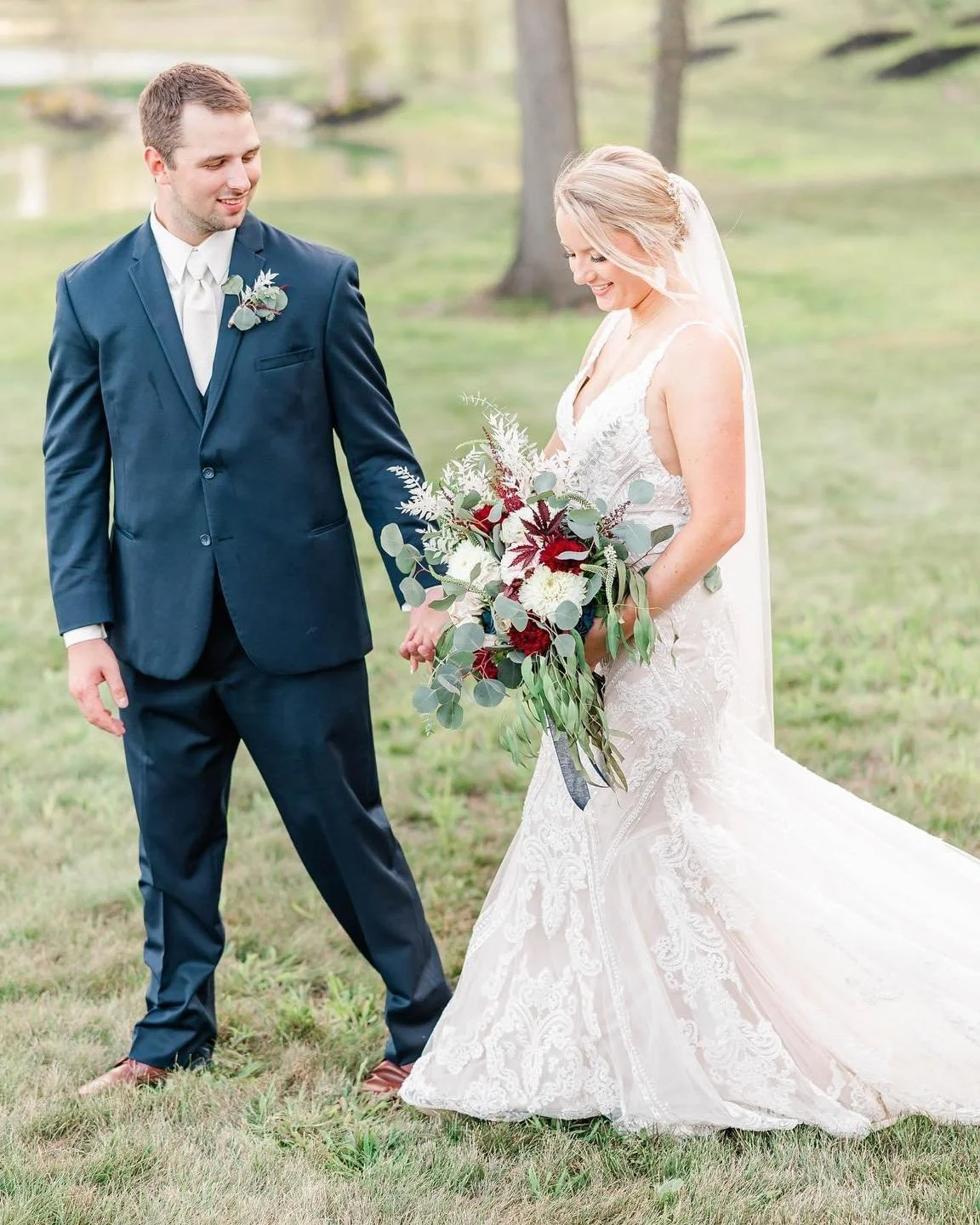 A bride and groom standing outdoors on a grassy area with trees in the background, holding hands and smiling at each other. The bride is wearing a white lace wedding gown and veil, holding a large bouquet of flowers with red and white blooms and gree
