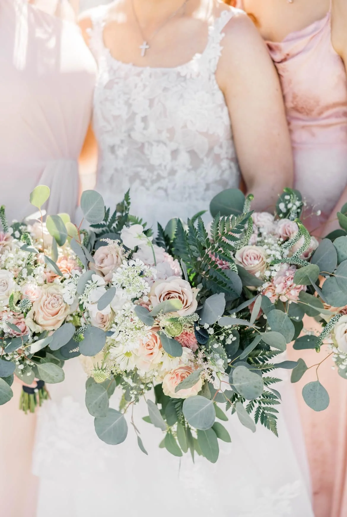 Close-up of a wedding bouquet with blush pink roses, white flowers, and various green foliage held by a bride wearing a detailed lace wedding dress and a cross necklace.