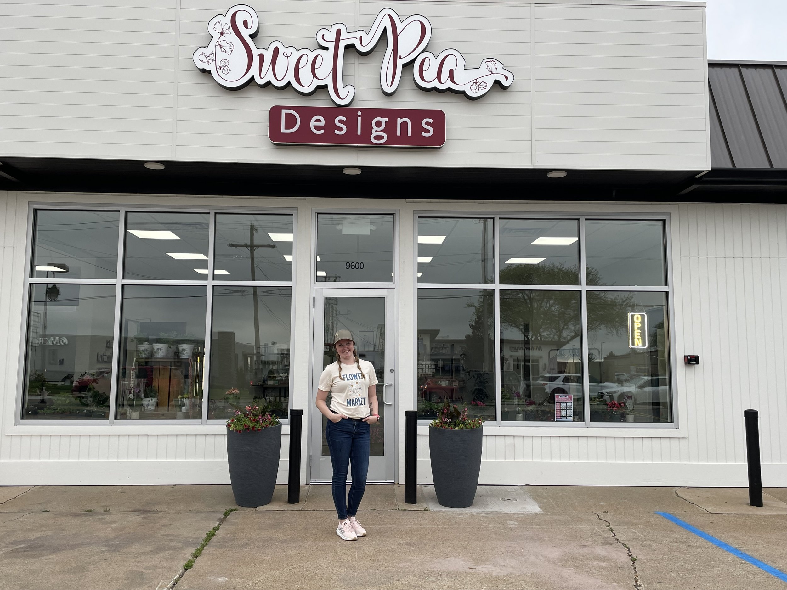 A woman standing in front of a storefront called 'Sweet Pea Designs' with large windows and potted flowers on either side of the entrance, on an overcast day.