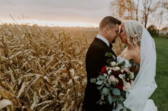 Bride and groom standing close with their foreheads touching in a field of dried corn stalks at sunset, the bride holding a bouquet of flowers, wearing a white wedding gown and veil, and the groom in a black suit.