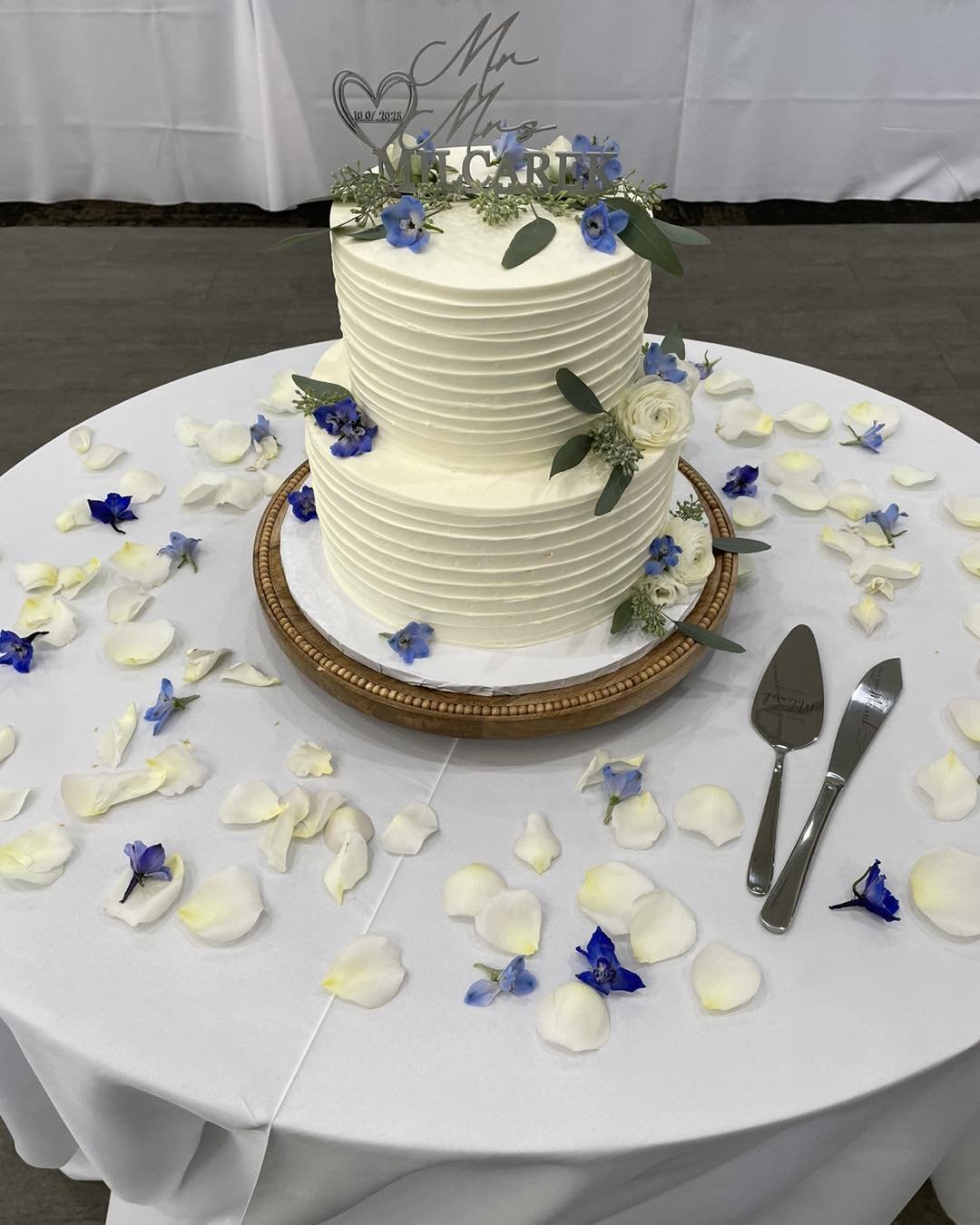 Two-tier white wedding cake decorated with blue flowers and green leaves on a round wooden cake stand, surrounded by scattered white and blue flower petals on a white tablecloth, with cake serving utensils nearby.