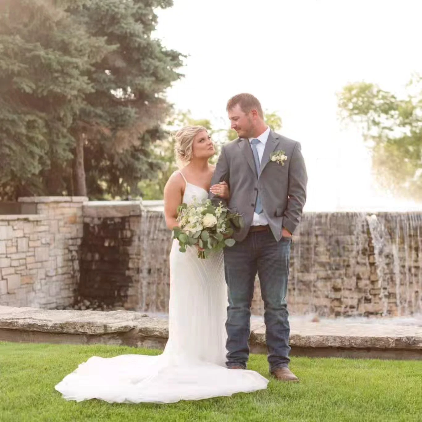 Bride and groom standing outside near a stone waterfall fountain, the bride holding a bouquet of white flowers and greenery, the groom dressed in a gray suit with a boutonniere, gazing at each other in a romantic moment during their wedding.