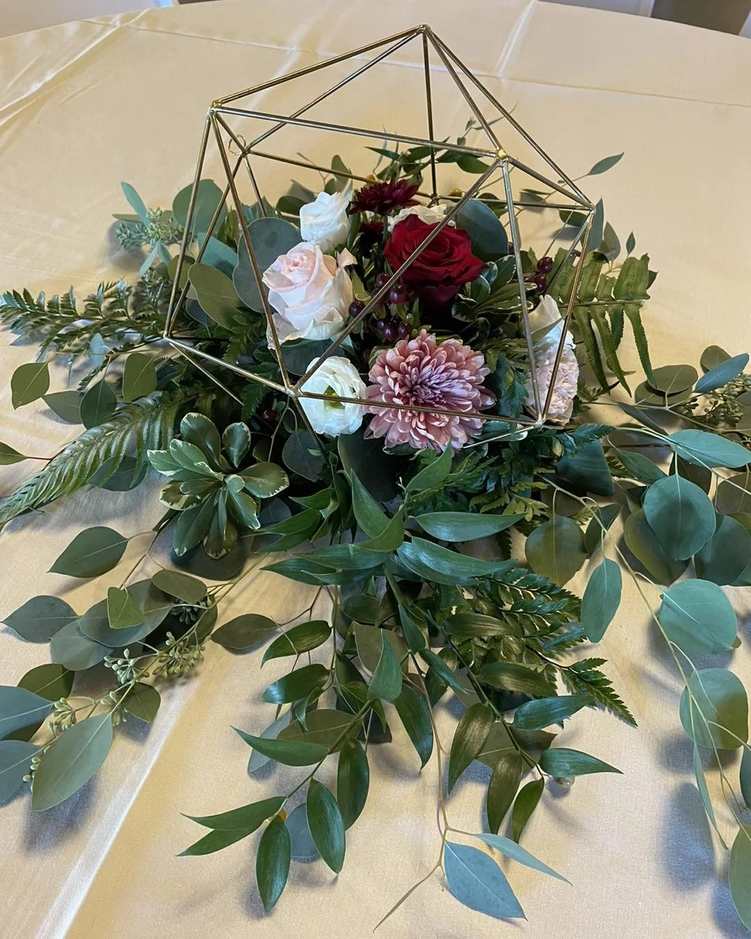A floral centerpiece with pink, white, and red roses, pink chrysanthemum, and greenery arranged beneath a geometric gold metal sculpture on a cream-colored tablecloth.