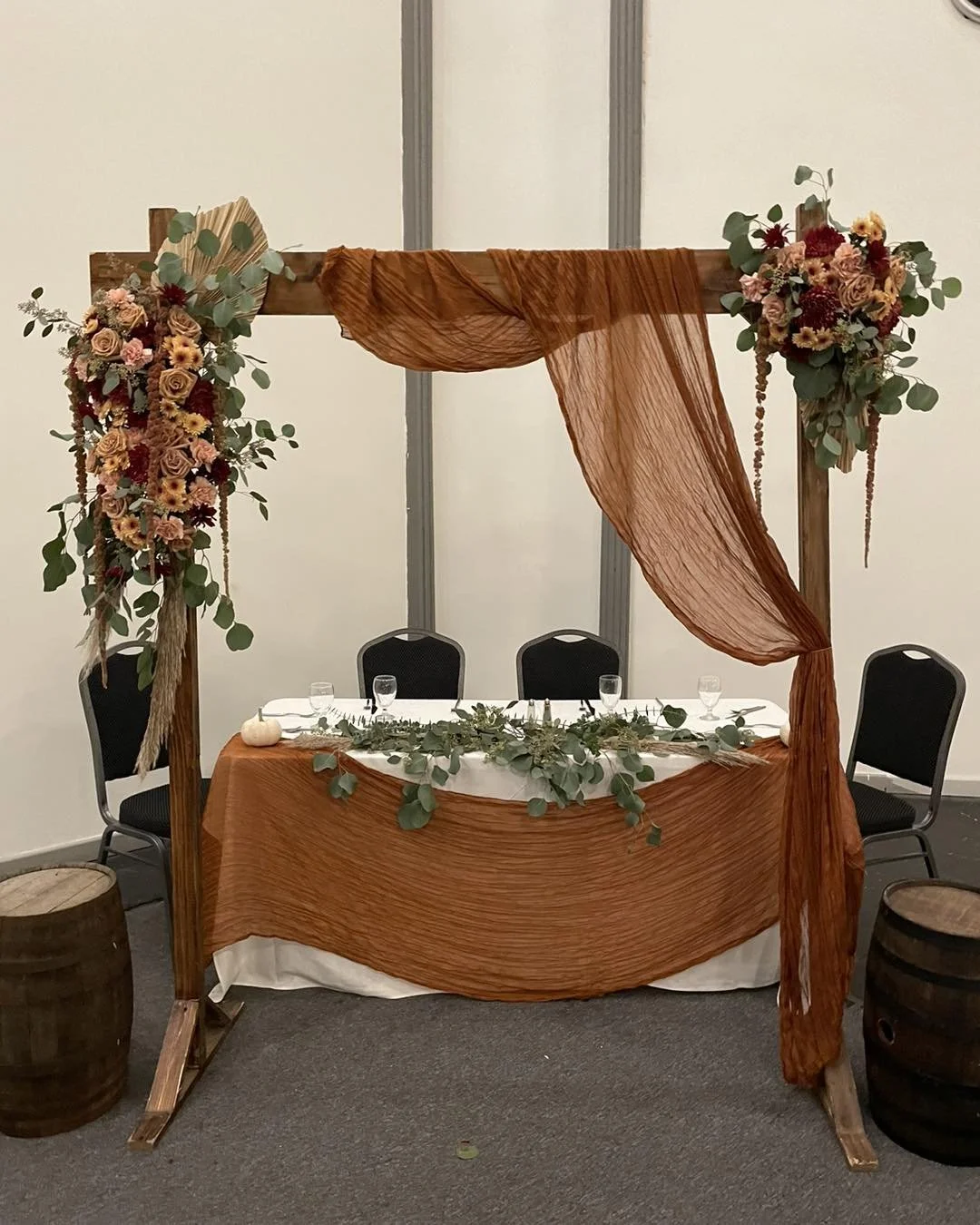 Rust-colored fabric draped over a wooden frame decorated with floral arrangements on top and greenery on the table. The setup appears to be for a wedding or celebration event.