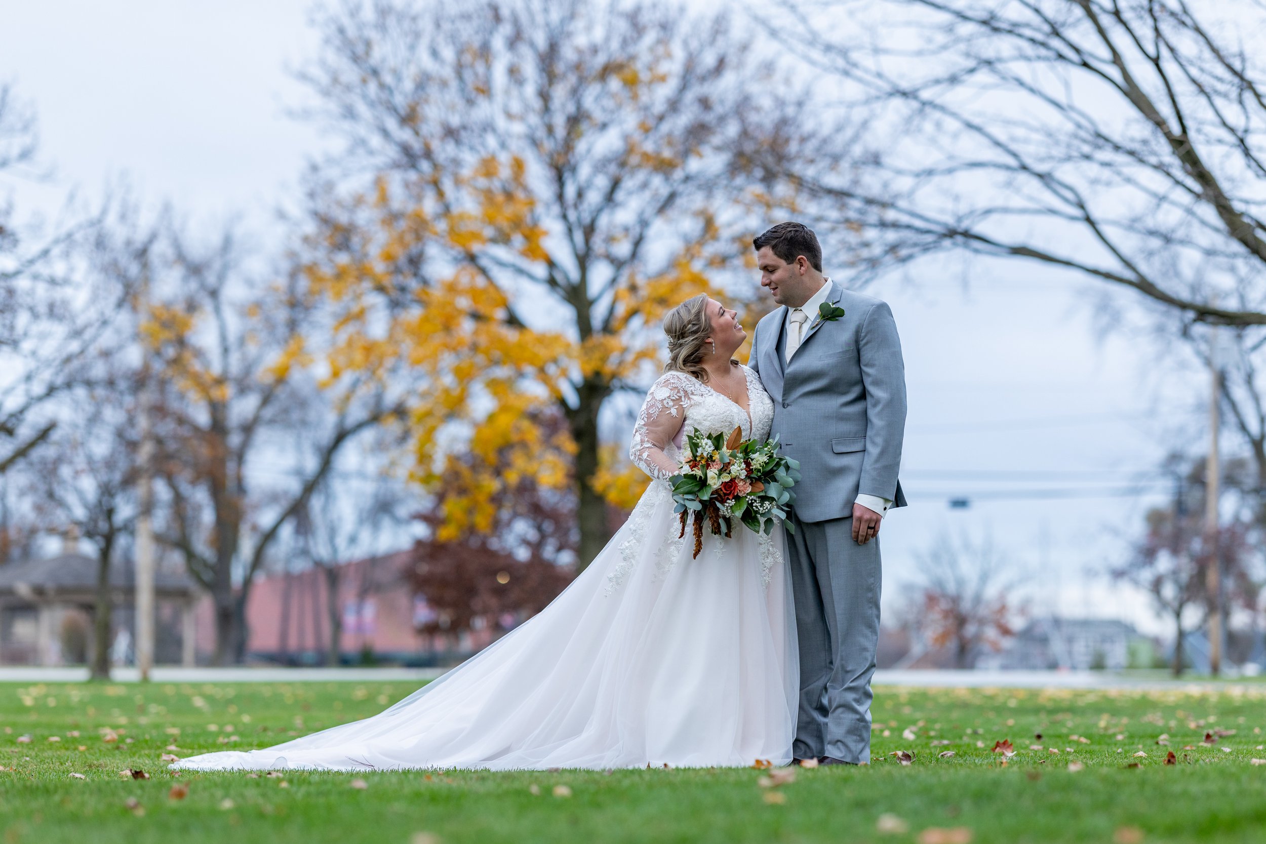 A bride and groom on their wedding day standing on grass, with trees and autumn leaves in the background. The bride wears a white lace wedding gown and holds a bouquet, while the groom wears a light gray suit and has a boutonniere.