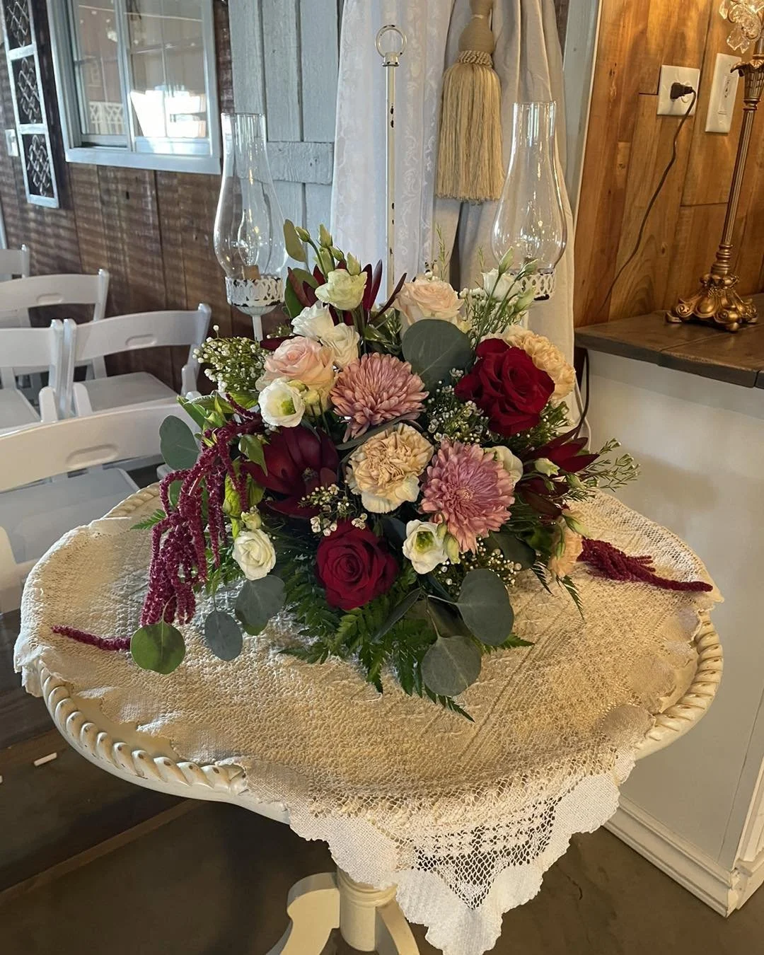 A floral arrangement with pink, white, and red roses, pink dahlias, white lisianthus, and greenery on a lace-covered round table in a cozy, rustic room.