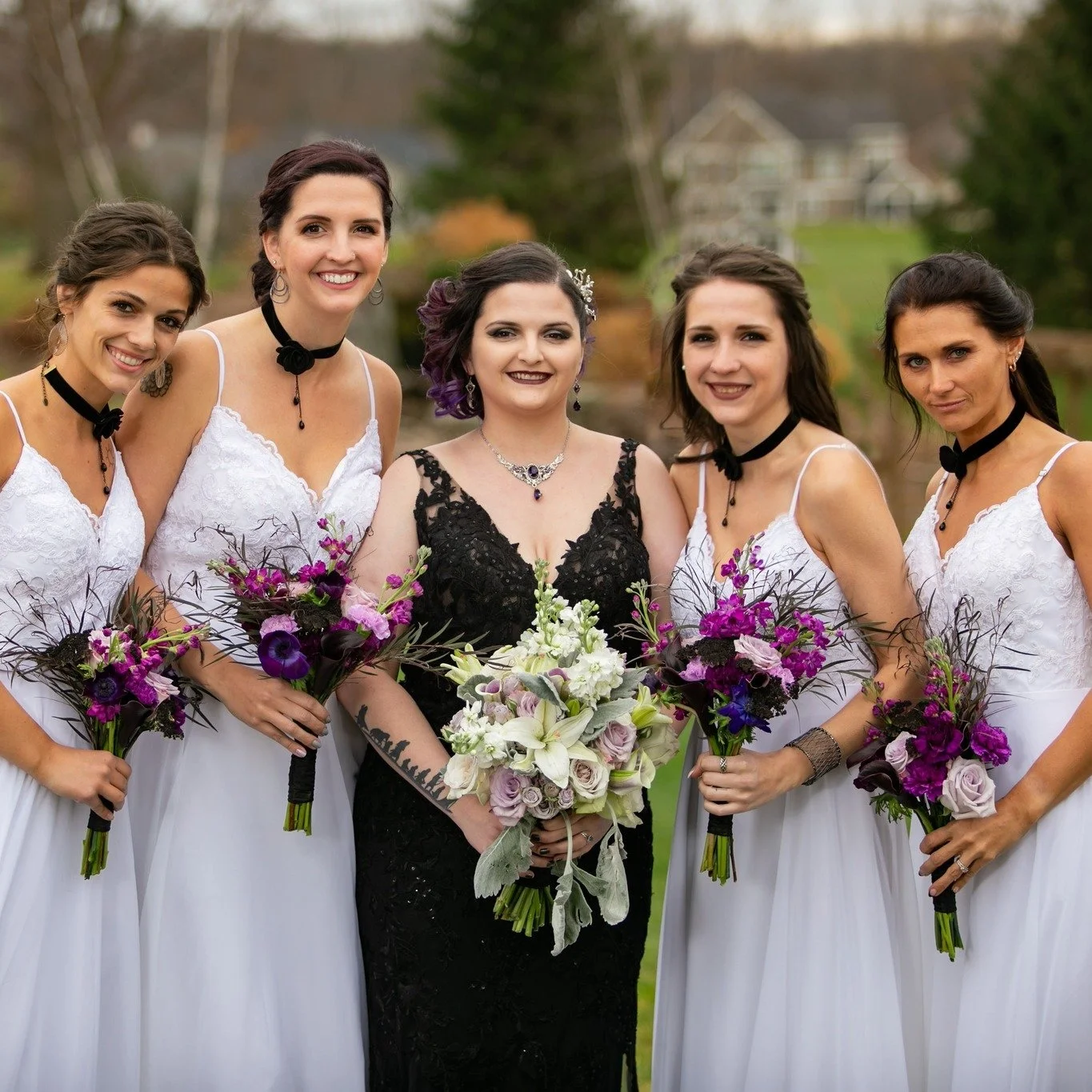 Group of six women in wedding dresses and a black dress outdoors holding bouquets.