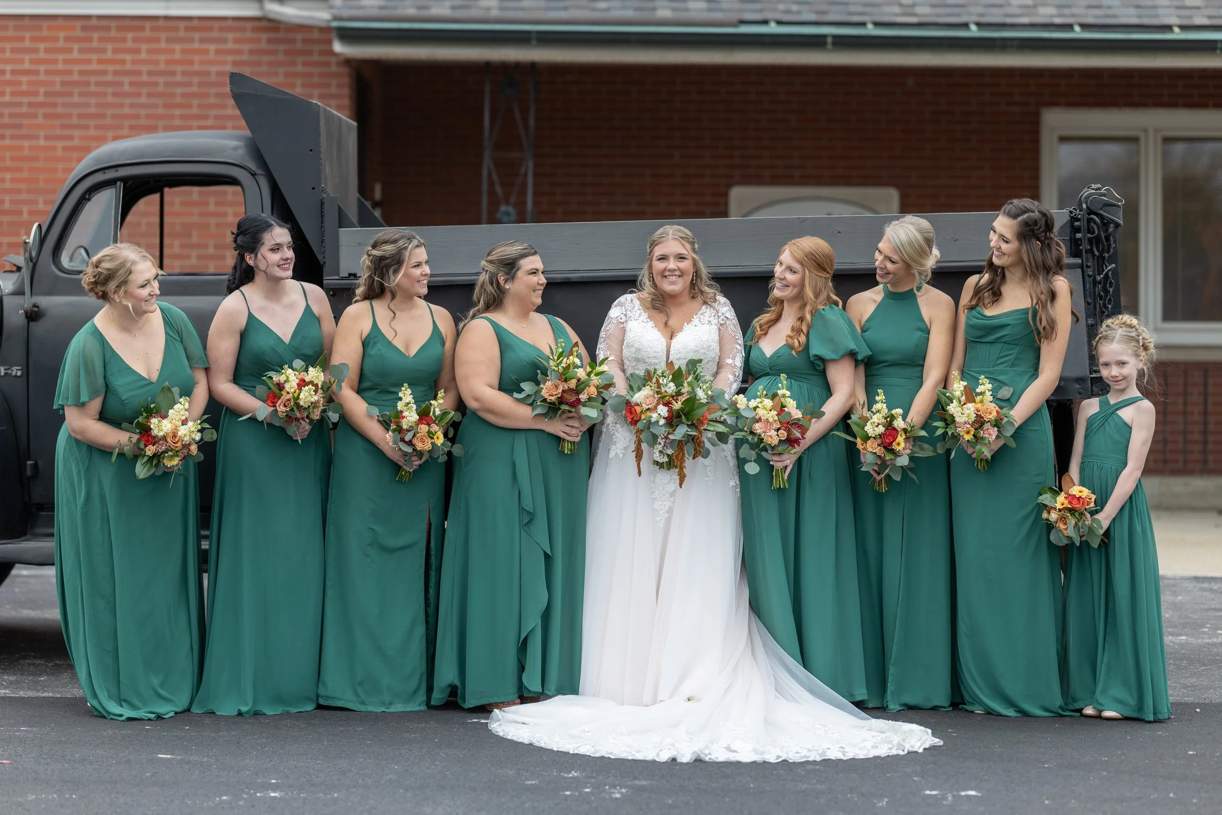 Bridal party standing in front of a black vintage truck on a wedding day, wearing green dresses and holding floral bouquets.