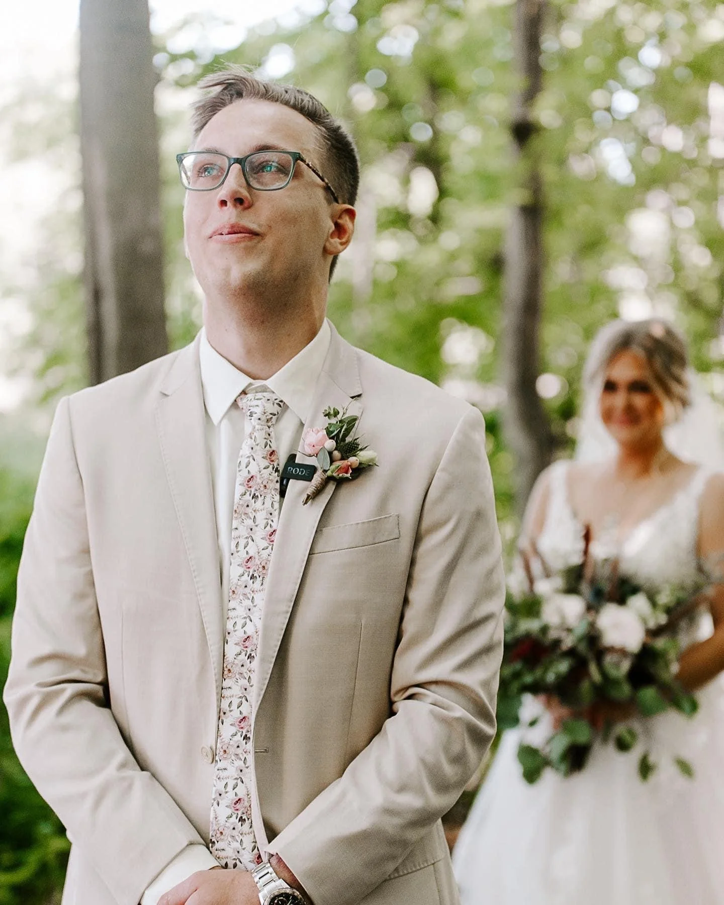 Groom in a beige suit and glasses standing outdoors with a bride in a white wedding dress holding a bouquet in the background.
