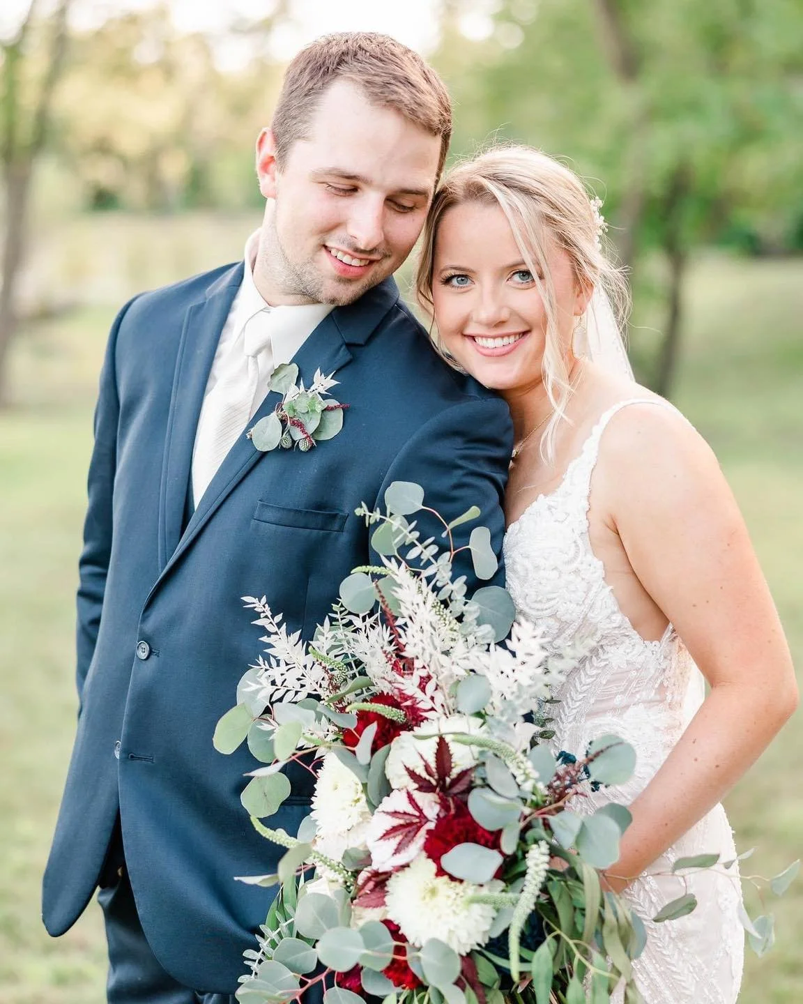 A newlywed couple outdoors, smiling and embracing, with the bride holding a large bouquet of flowers and greenery.