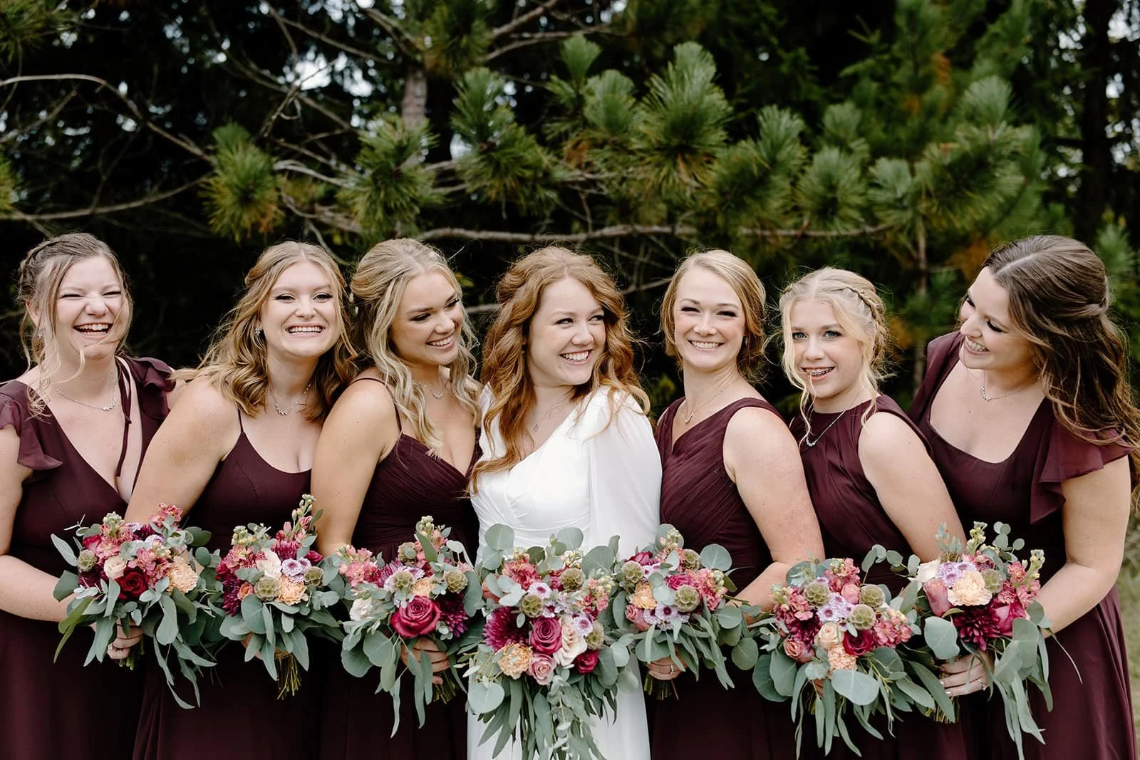 Bride with six bridesmaids outdoors, holding floral bouquets, smiling, with green trees in the background.