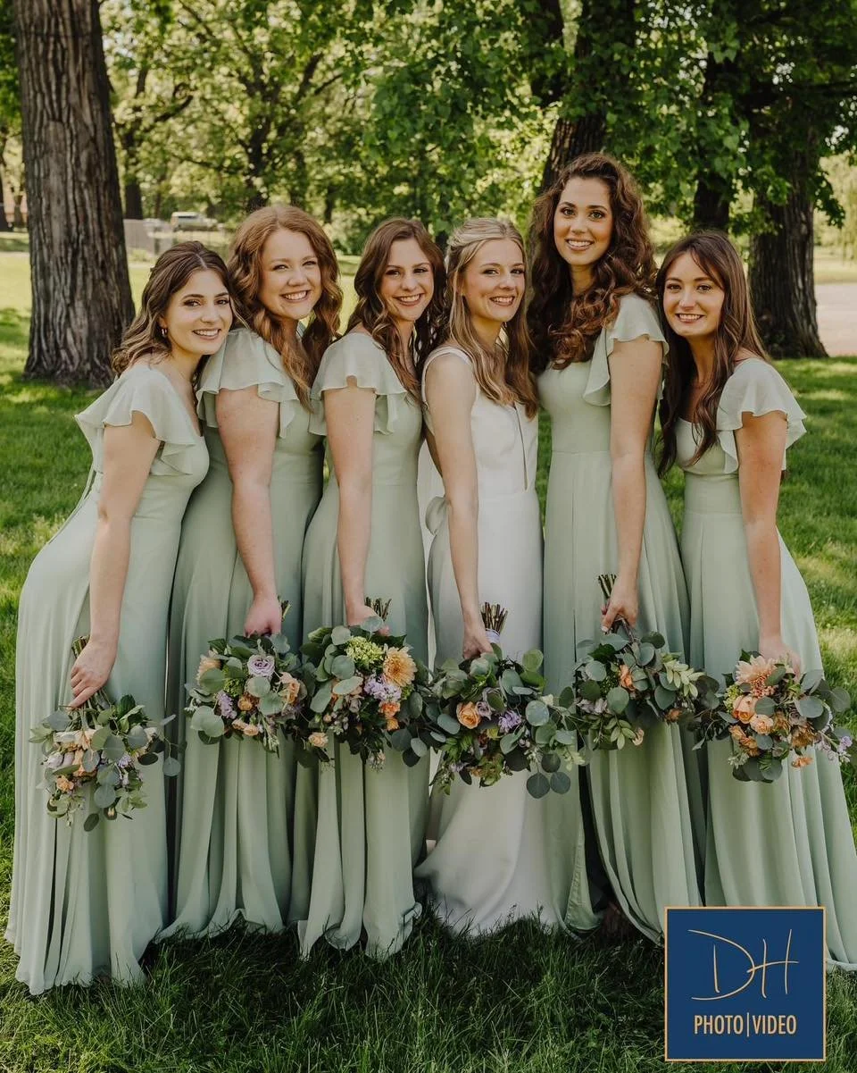 Seven women in matching light green dresses standing outdoors on grass with trees, holding bouquets of flowers, smiling for a group photo.