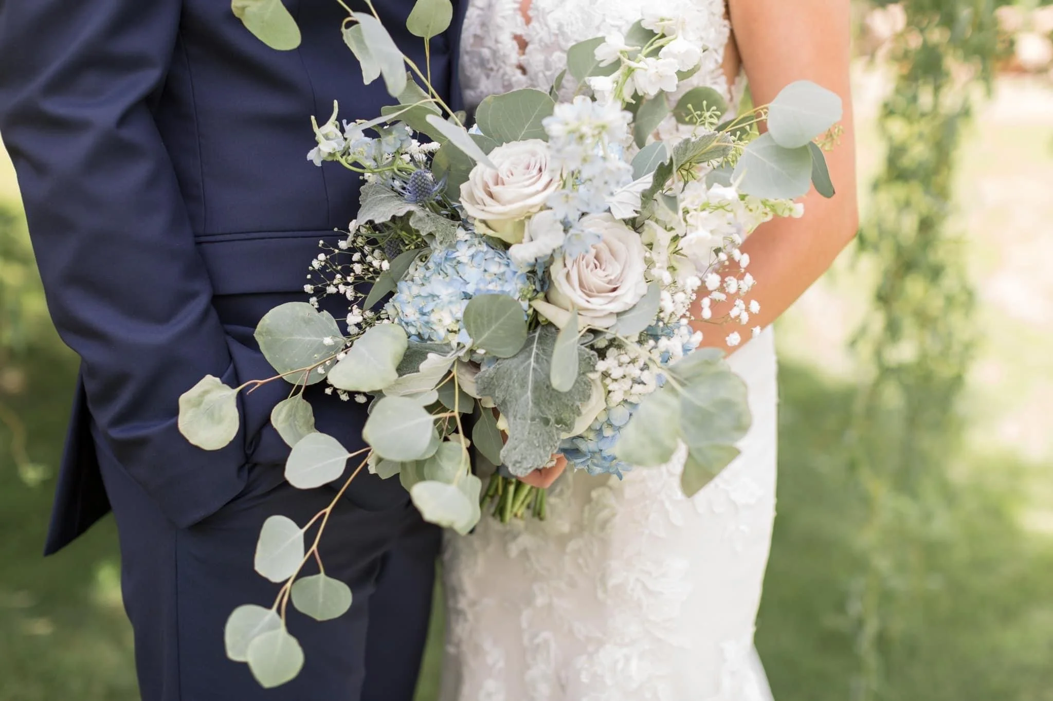 Close-up of a bride and groom holding a bouquet of white roses, blue hydrangeas, and greenery outdoors.