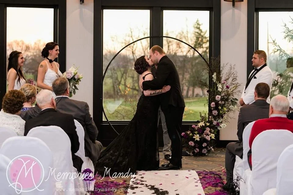 A couple shares a kiss during their wedding ceremony in front of a large window, with guests seated on white chairs and flower arrangements nearby.