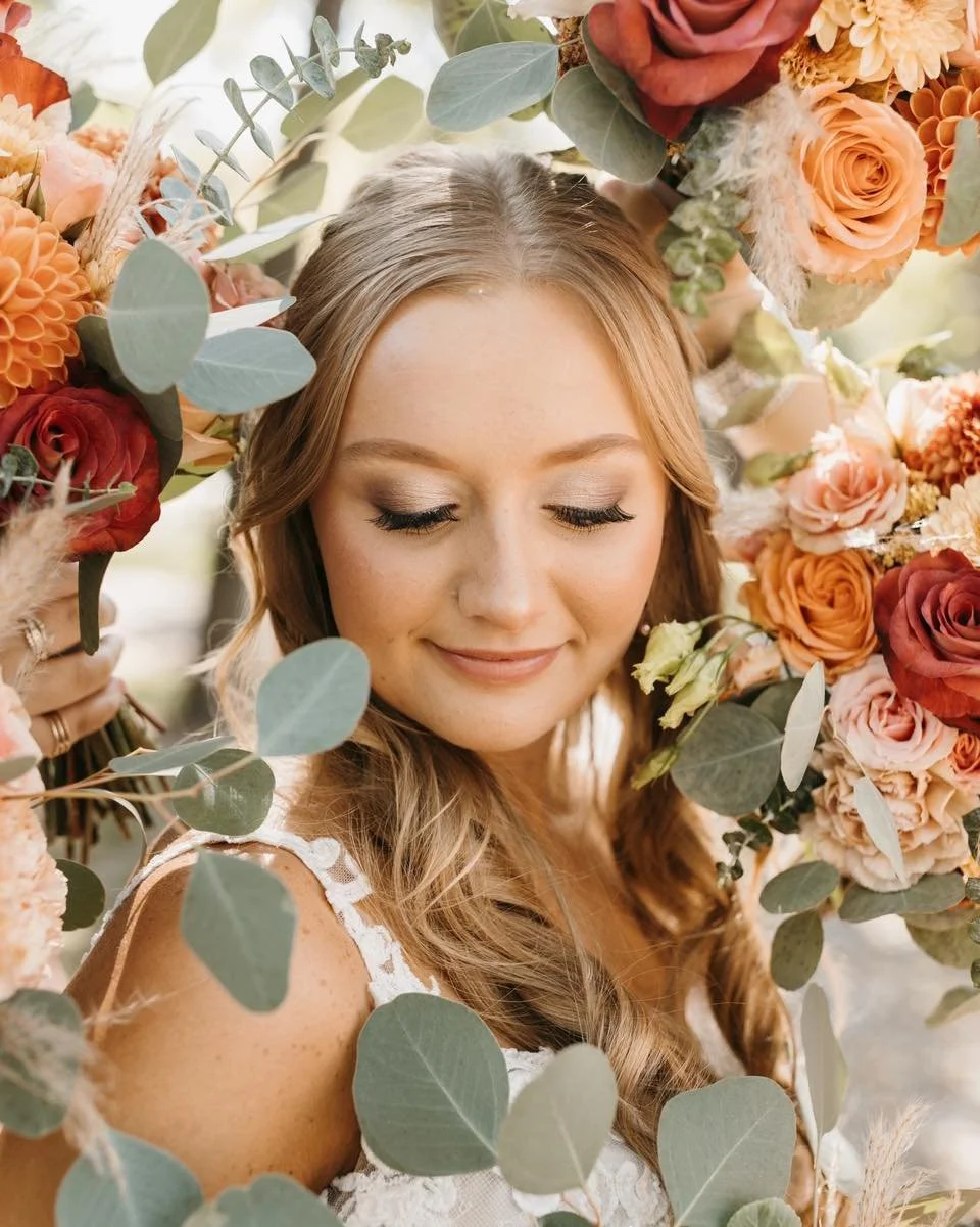 A woman with long, wavy blonde hair and closed eyes, smiling softly, surrounded by a large wreath of peach, pink, and orange roses, dahlias, and eucalyptus leaves.