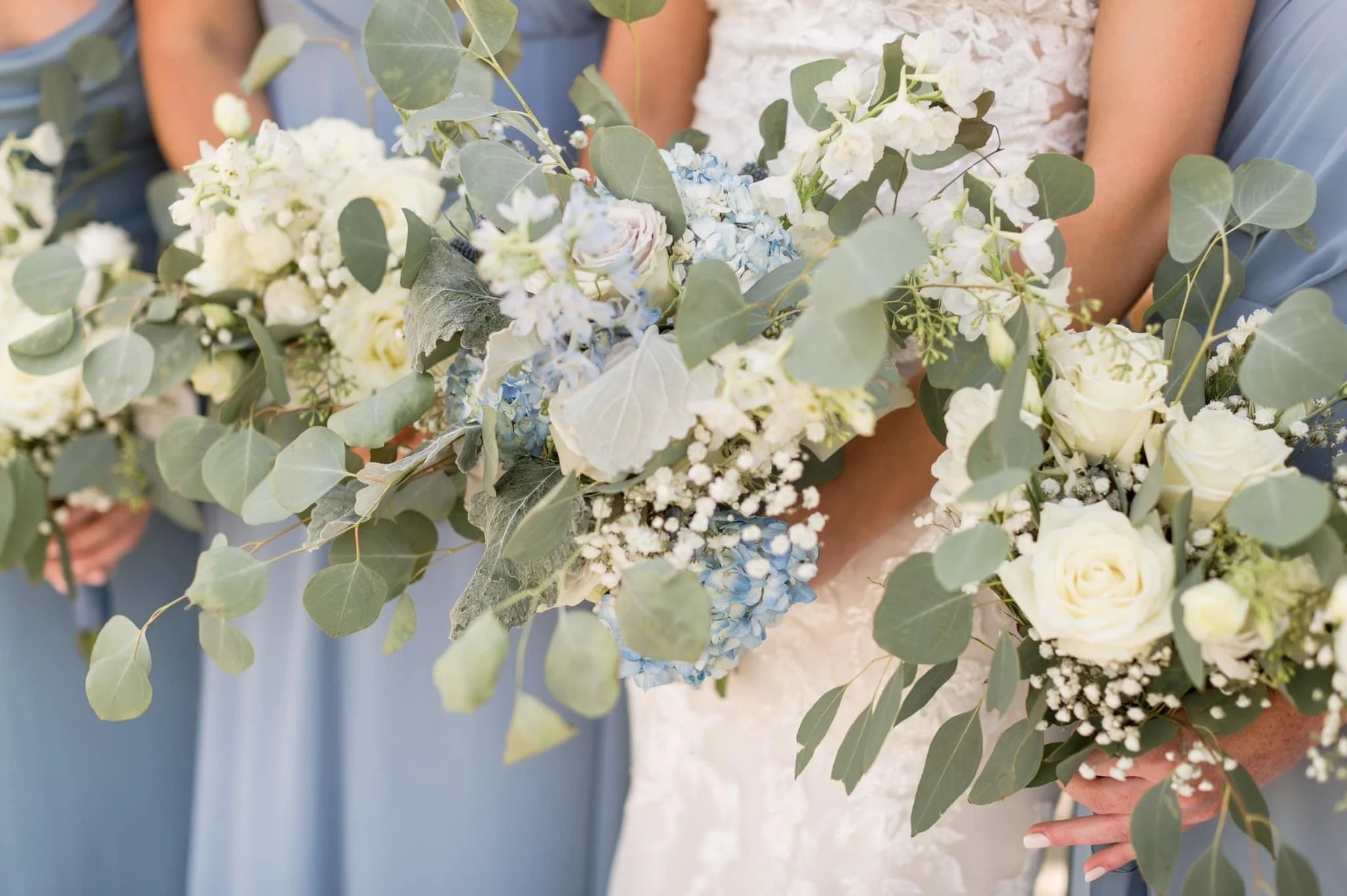 Close-up of a wedding bouquet featuring white roses, eucalyptus leaves, blue hydrangeas, and baby's breath, held by a bride in a lace dress with bridesmaids in light blue dresses in the background.