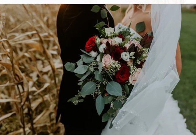 Bride in a white wedding dress holding a bouquet of red and pink roses, with greenery, standing next to a person in a black suit in an outdoor setting with dried plants and grass in the background.