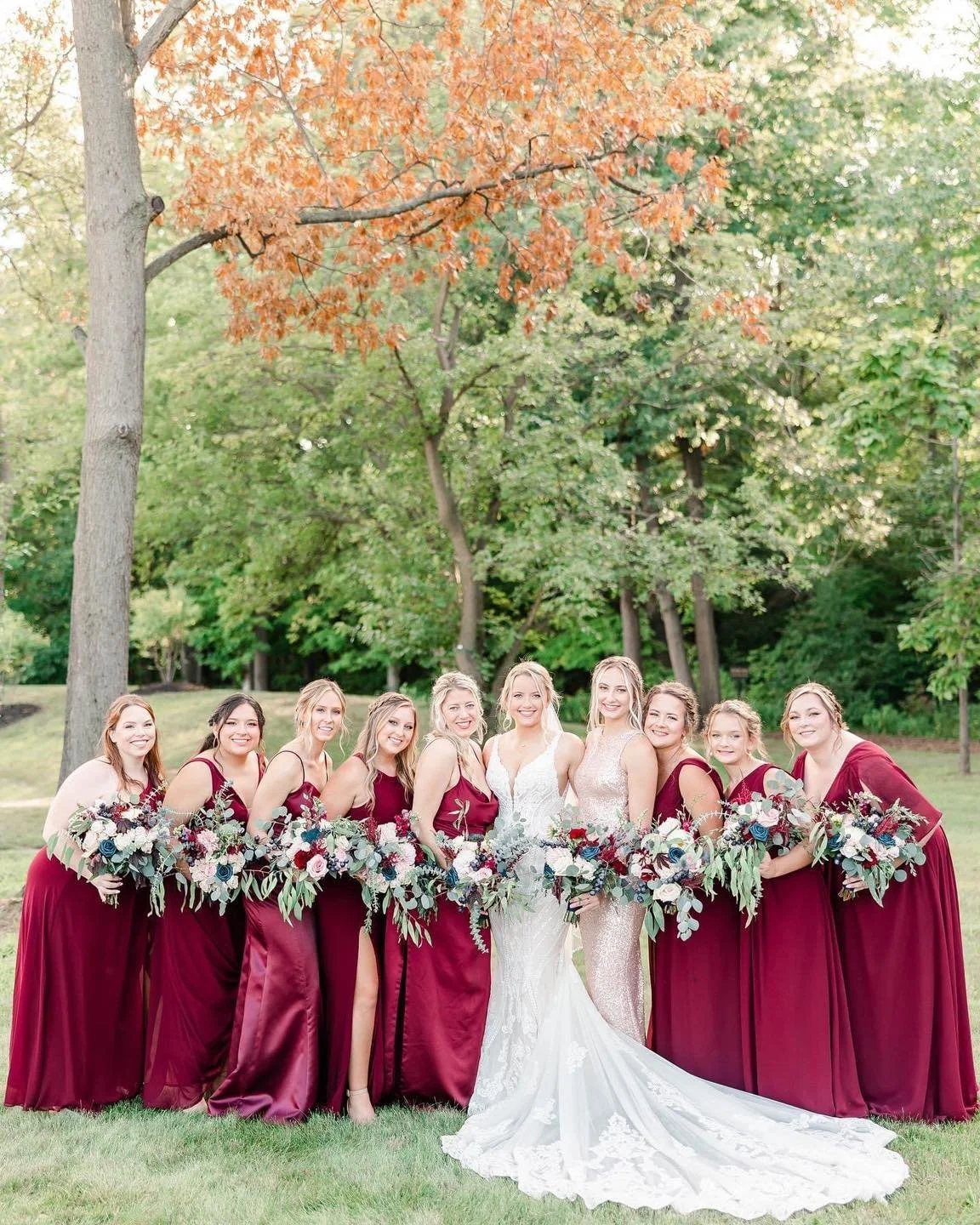 A group of women, including a bride in a white wedding gown, standing outdoors on grass with trees in the background, holding bouquets of flowers, during a wedding celebration.