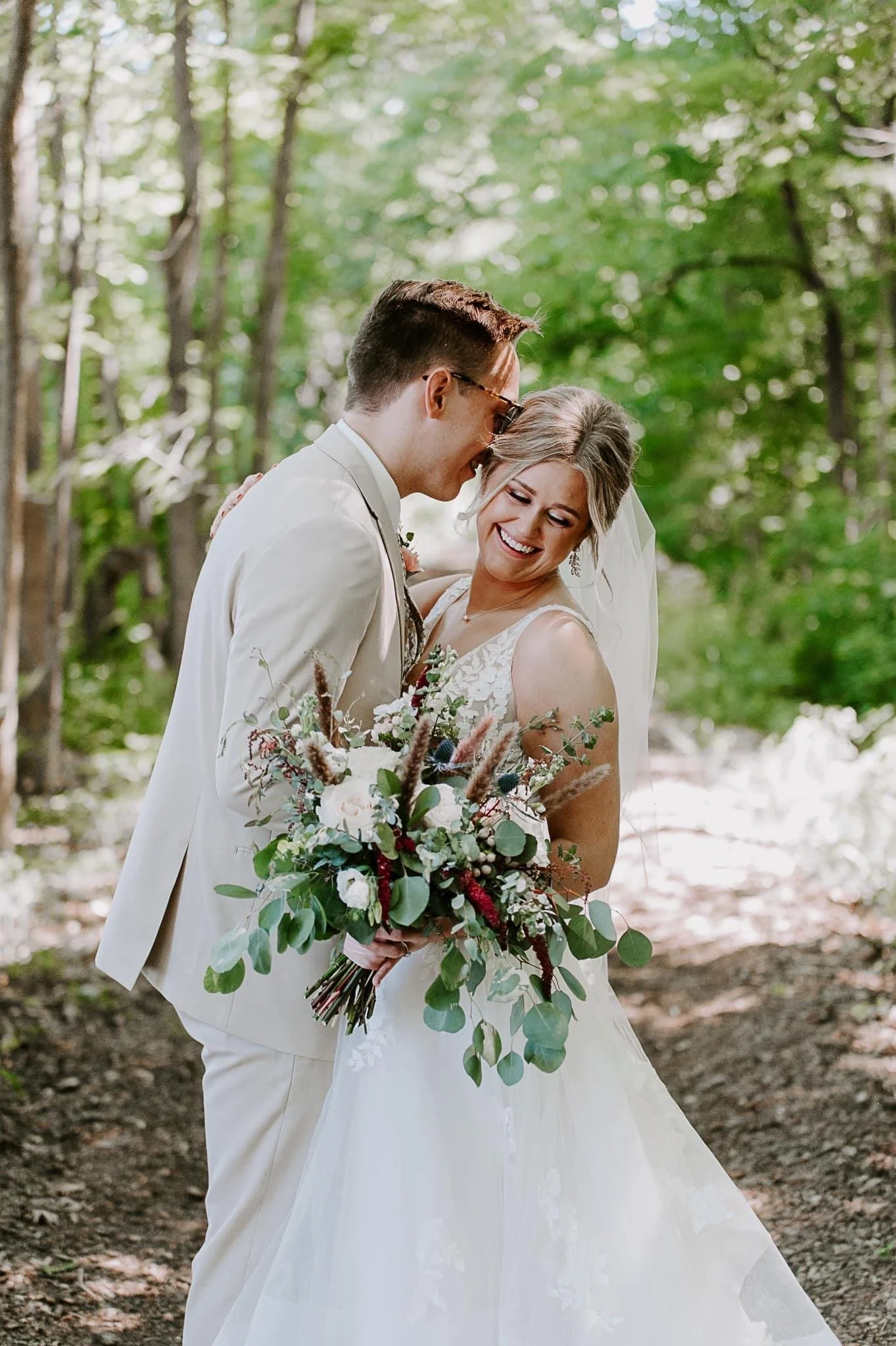 A bride and groom sharing a joyful moment outdoors, with the groom leaning his forehead against the bride's temple. The bride holds a large bouquet of flowers, and both are smiling, surrounded by green trees.
