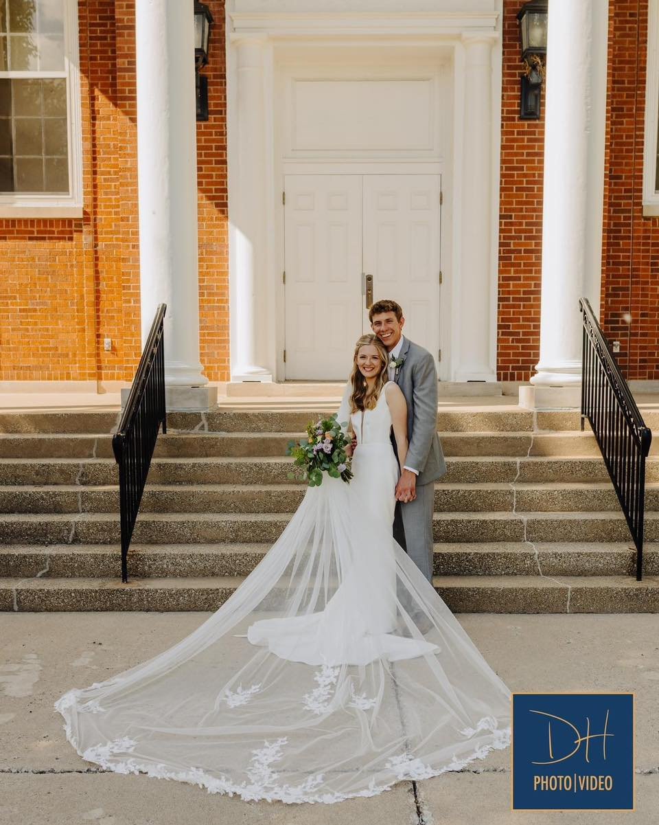 A newly married couple stands on the steps of a brick building with white columns, smiling. The bride is in a white wedding dress with a long veil and holding a bouquet of flowers. The groom is in a light gray suit.