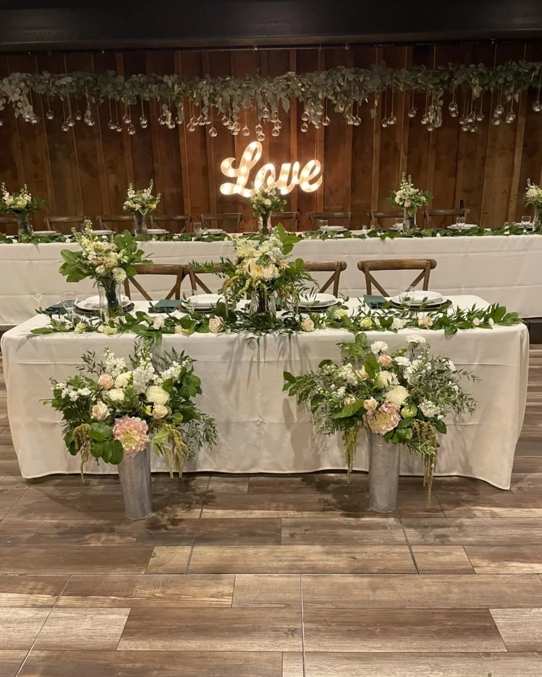 Wedding reception table decorated with white tablecloths, floral centerpieces, and greenery. A wooden backdrop features a lit sign spelling 'Love.' Hanging glass decorations and greenery adorn the ceiling.