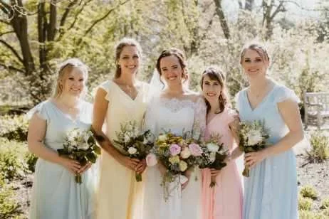 Group of five women,  a bride and bridesmaids, standing outdoors in a sunny garden with trees, all holding bouquets of flowers and dressed in pastel-colored dresses.