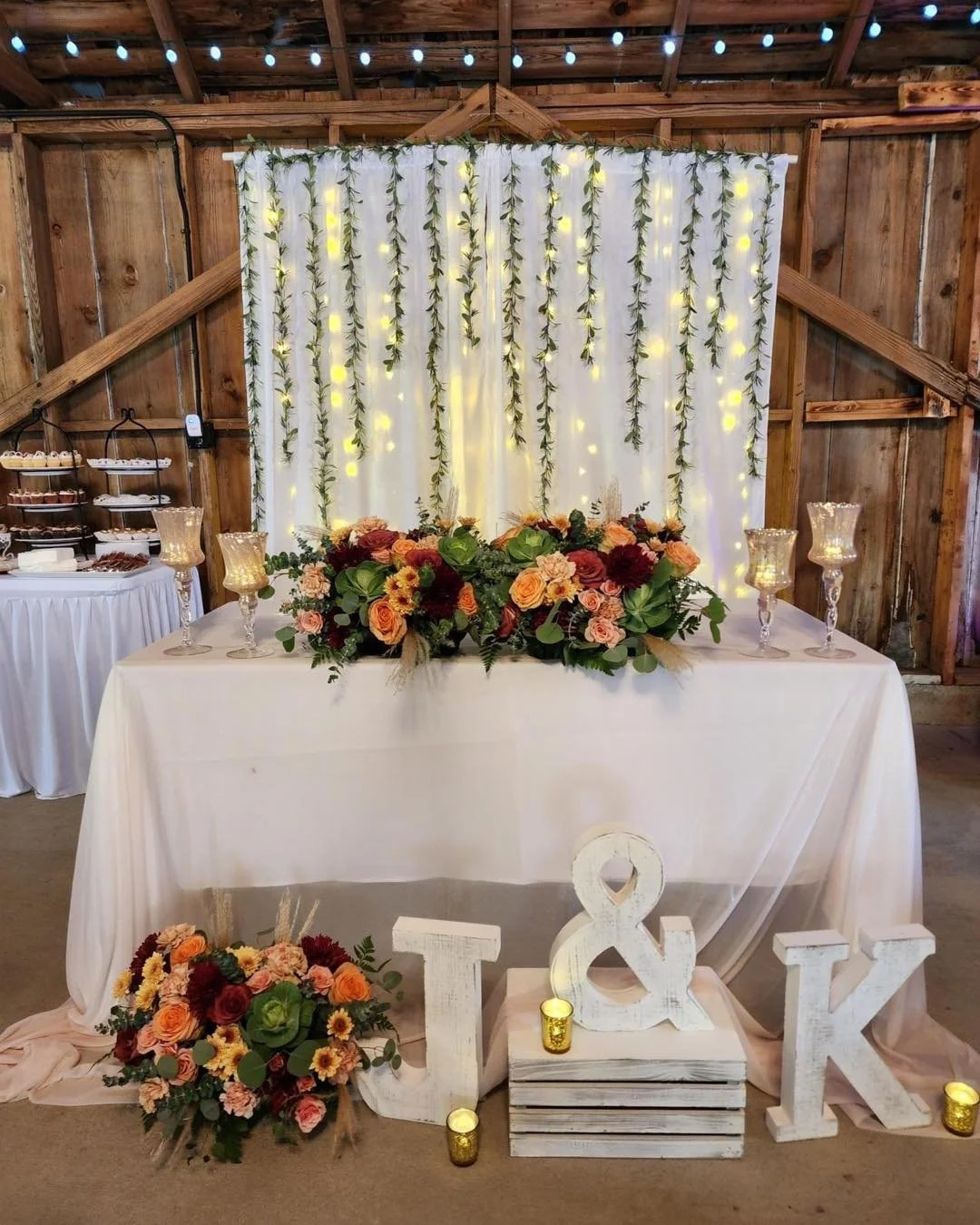 Decorated sweetheart table with floral arrangements, candles, and large white letters J & K in front, set in a rustic barn with string lights and a white backdrop of draped fabric and hanging greenery.