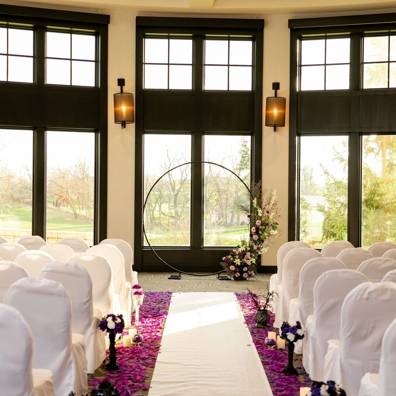 Indoor wedding ceremony setup with white covered chairs, floral arrangements, candles, and a circular metal arch at the front near large windows.