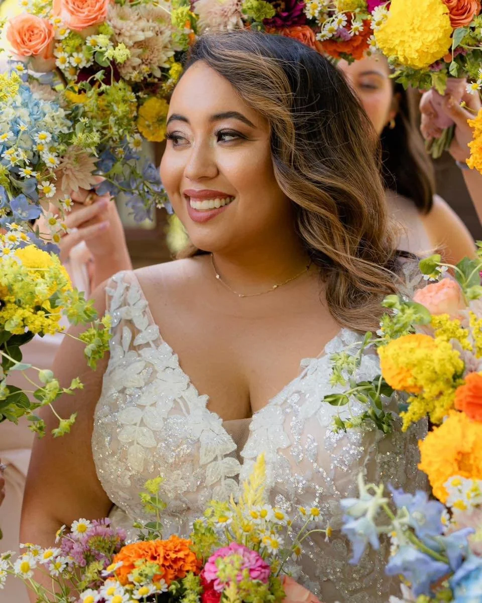 Woman in an elegant dress smiling among a colorful arrangement of flowers, with other women partially visible behind her.