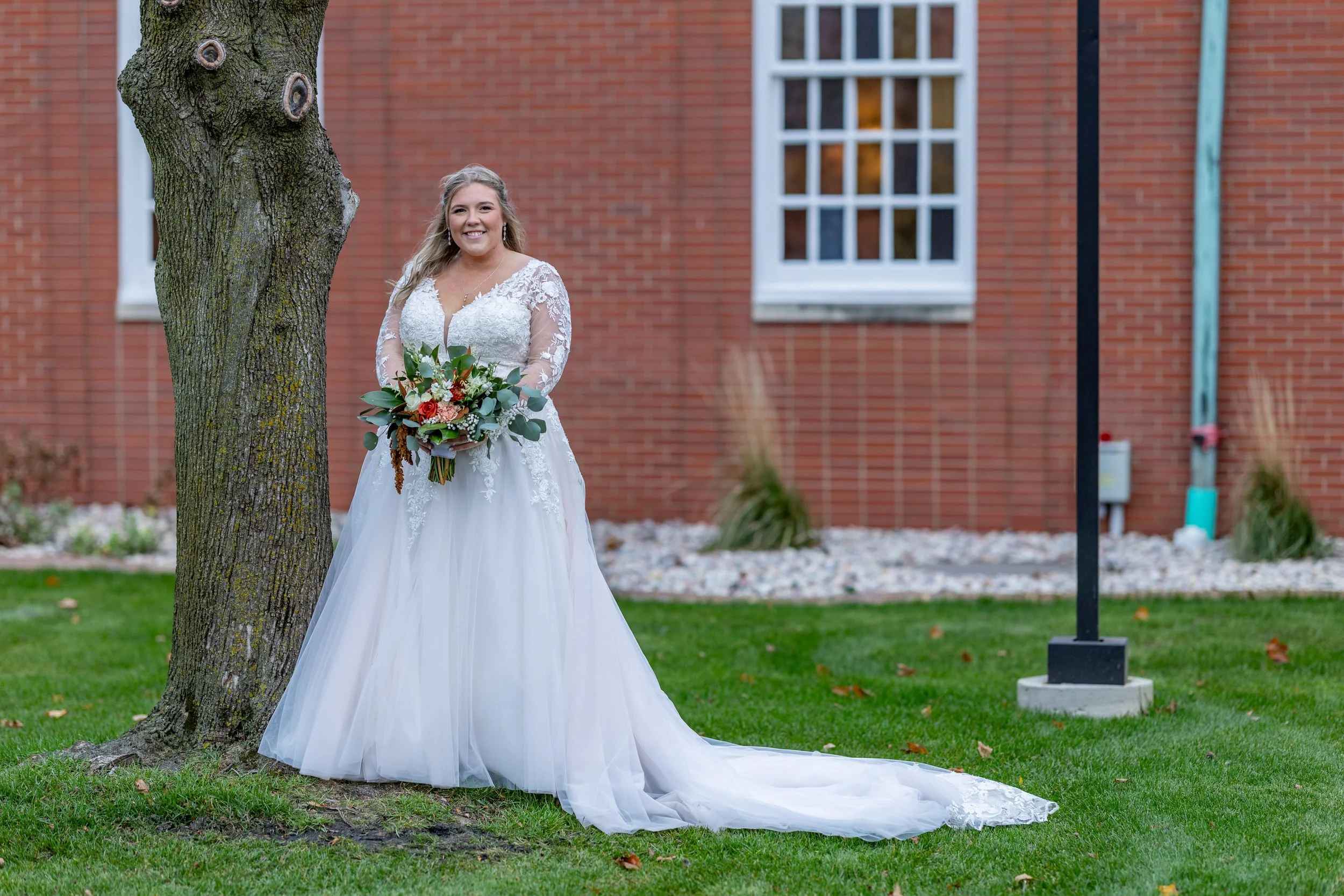 A bride in a white wedding dress smiling and holding a bouquet, standing outside on green grass near a tree and brick building.