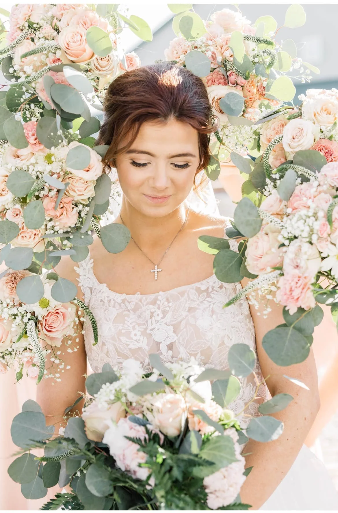Bride holding a bouquet of light pink and white flowers in front of her, standing among a large floral display of pink roses, white flowers, and green eucalyptus leaves, wearing a lace wedding dress with a cross necklace.