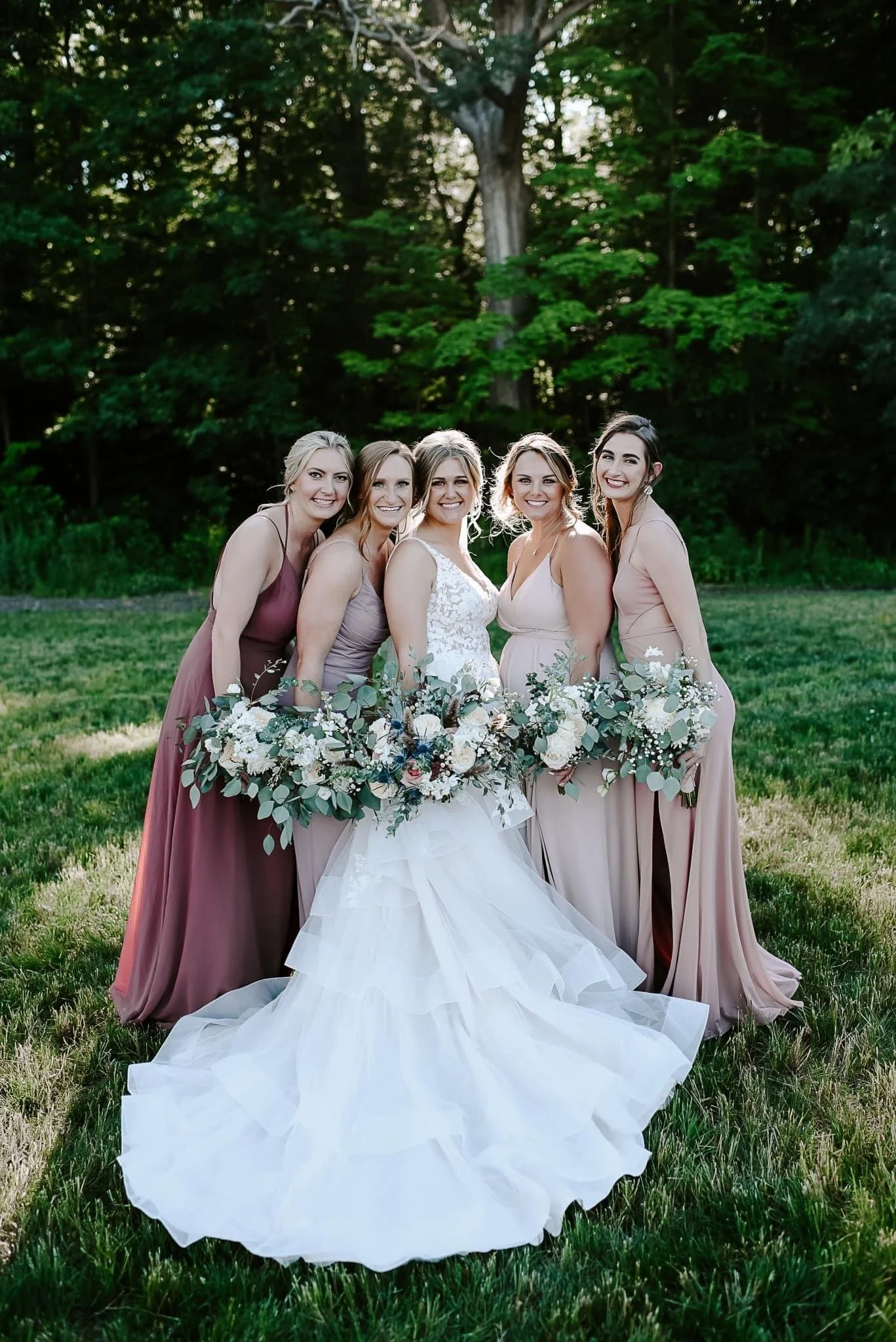 A bride and four bridesmaids standing together outdoors on grass, holding bouquets, with trees in the background. The bride is in a white wedding gown, and the bridesmaids are in mauve and blush dresses.