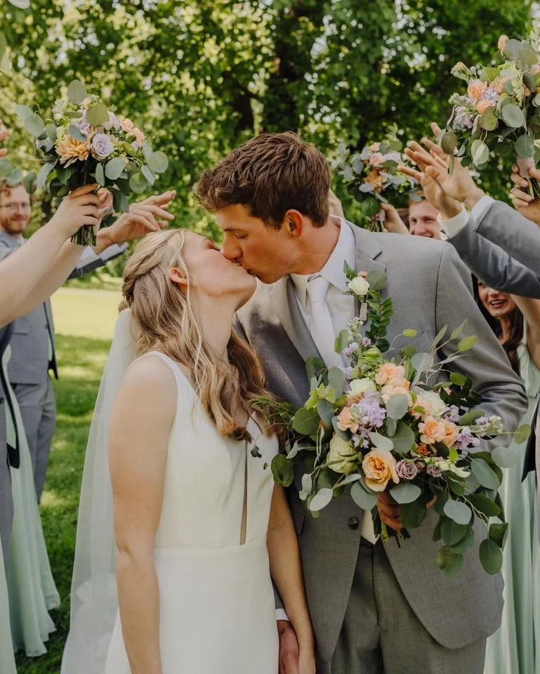 A bride and groom kiss during their outdoor wedding ceremony, surrounded by friends holding bouquets of flowers.
