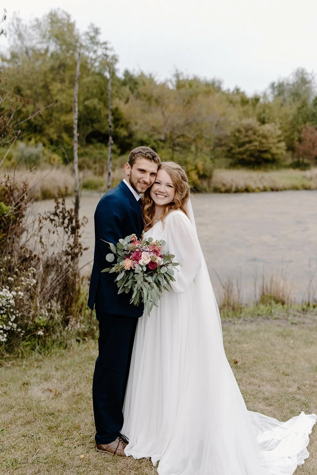 A newlywed couple stands smiling outdoors, the bride holding a bouquet of pink and red roses with greenery, embracing near a pond with trees and autumn foliage in the background.