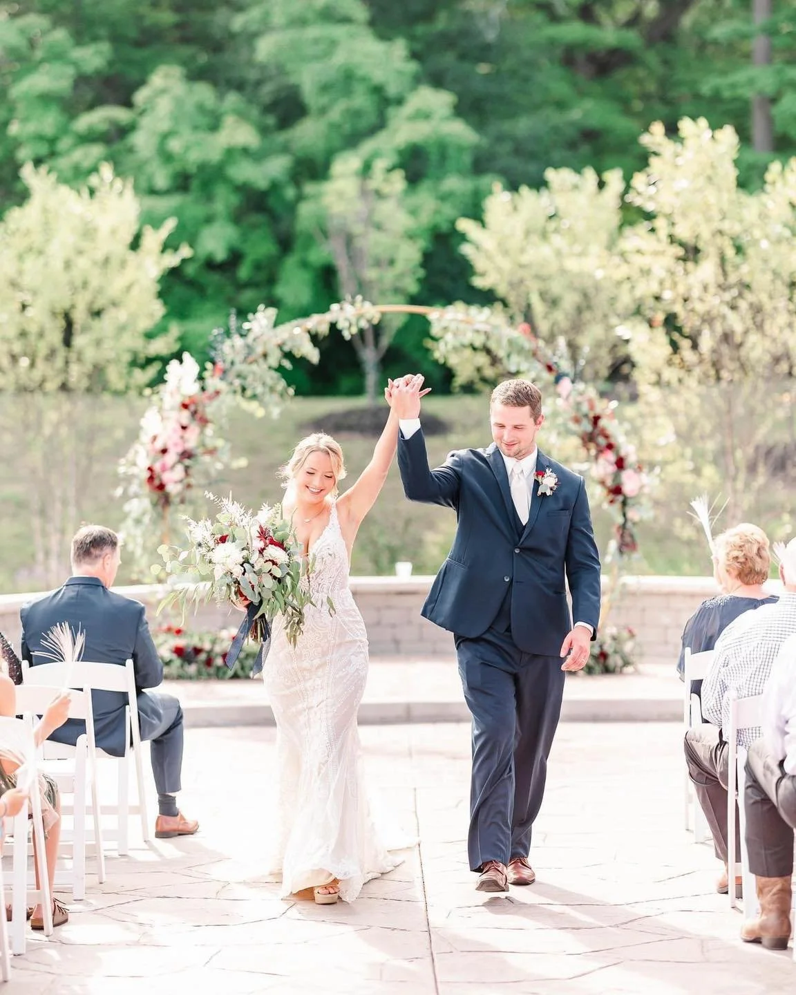 A newlywed couple walking down the aisle at their outdoor wedding ceremony, smiling and holding hands, with guests seated on either side and a floral arch in the background.