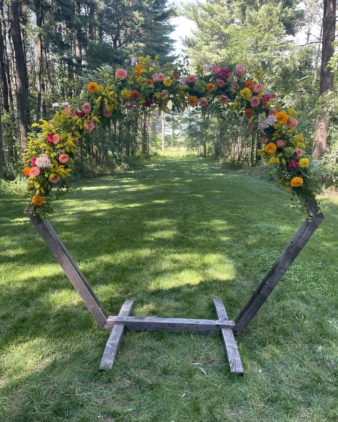 Wooden arch decorated with colorful flowers including pink roses, yellow daisies, and orange marigolds, set outdoors in a grassy, wooded area.
