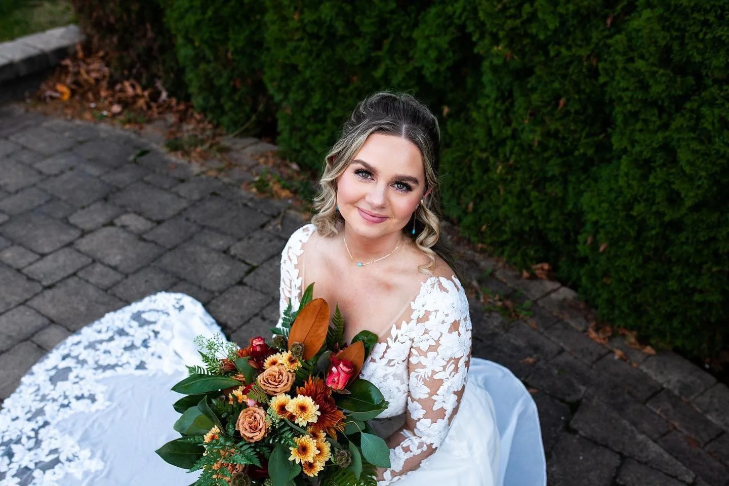 A bride with blonde curly hair and a lace wedding dress holding a vibrant bouquet of flowers looking up and smiling outdoors.