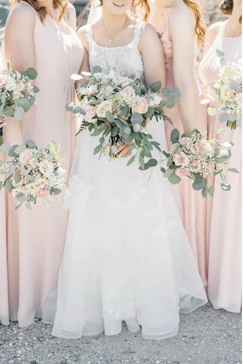 Bridal party with women in pink dresses holding bouquets of pink and white flowers with green foliage, standing outdoors on gravel ground.