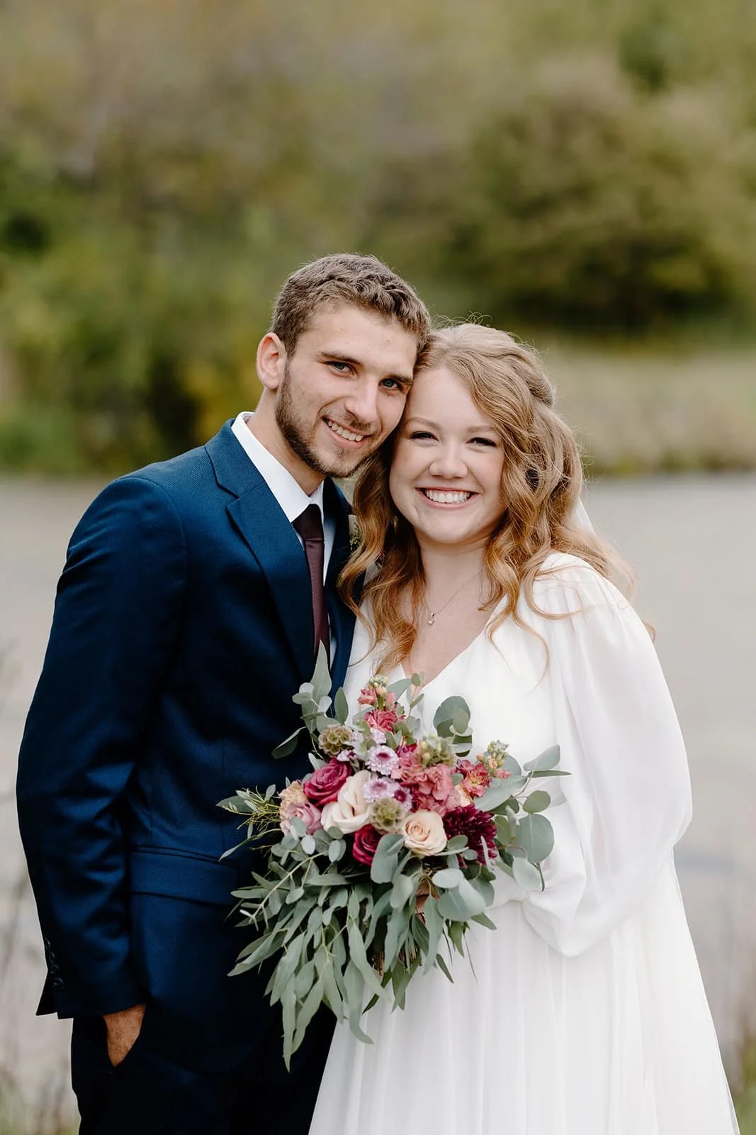 A smiling couple dressed in wedding attire outdoors, with the woman holding a bouquet of pink, white, and green flowers, standing in front of a blurred natural landscape.