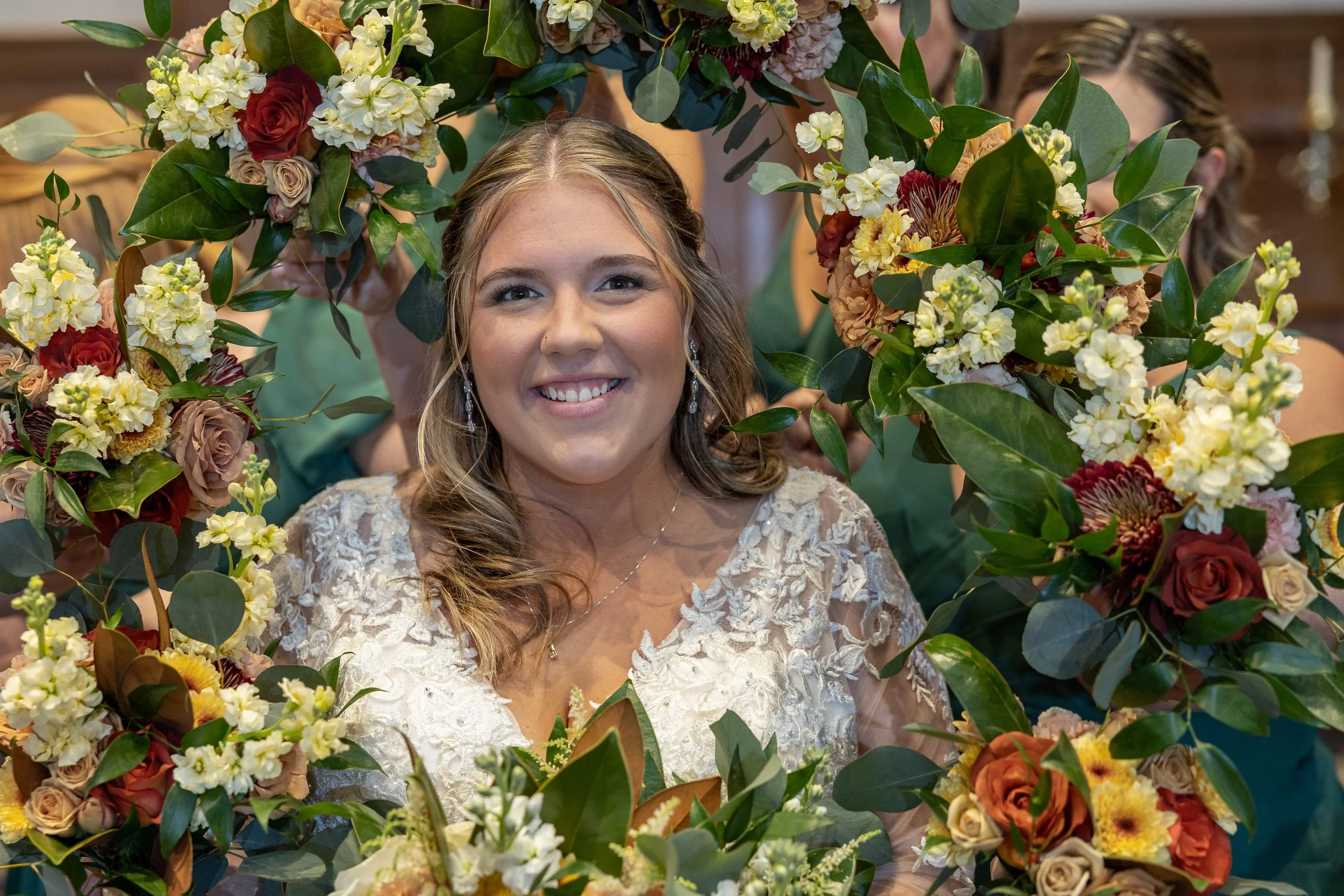 A smiling woman in a white lace dress is seen through a floral arch with various colorful flowers and green leaves.