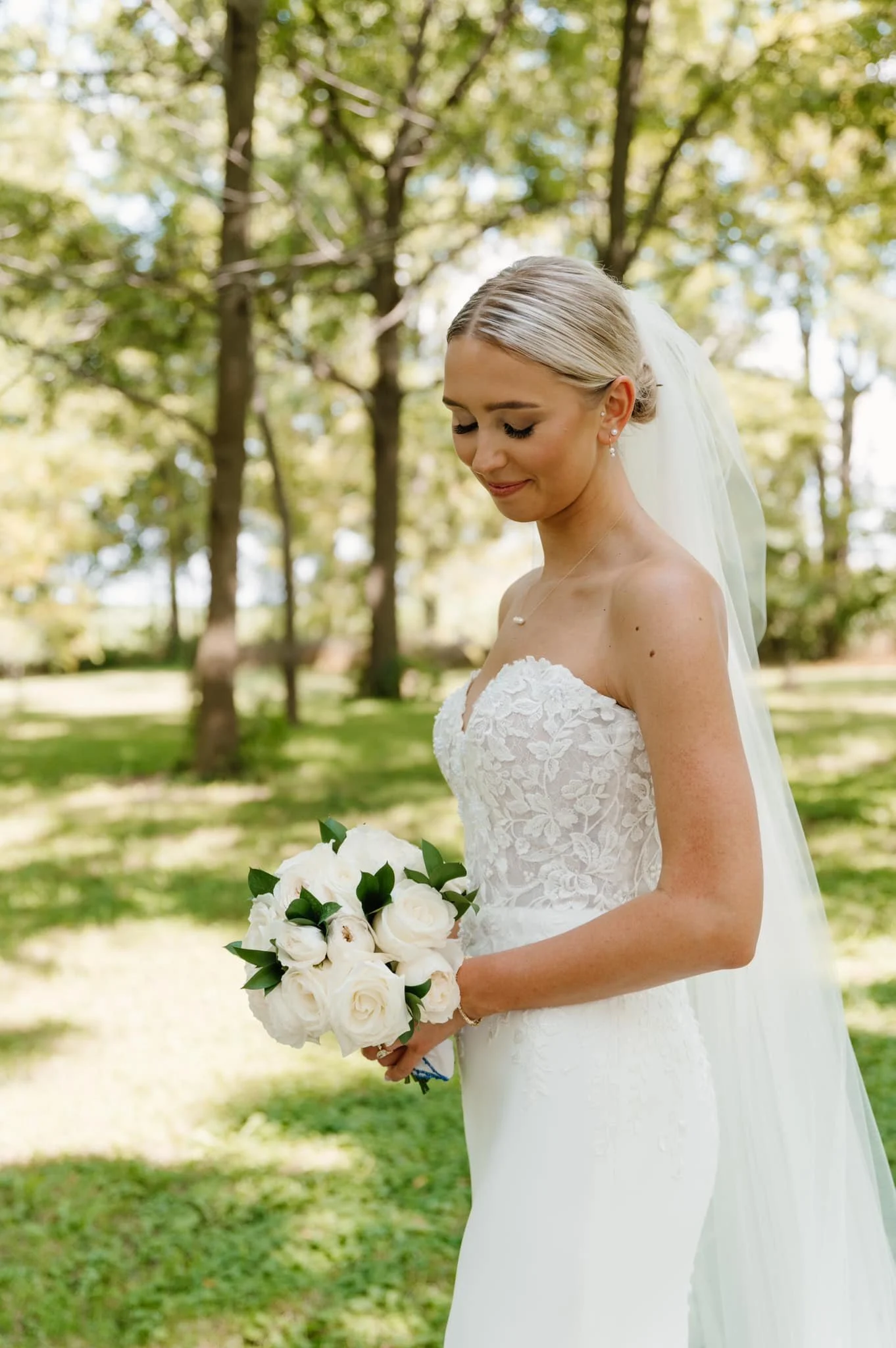 A bride in a white lace wedding dress holding a bouquet of white roses outdoors in a park with green trees.
